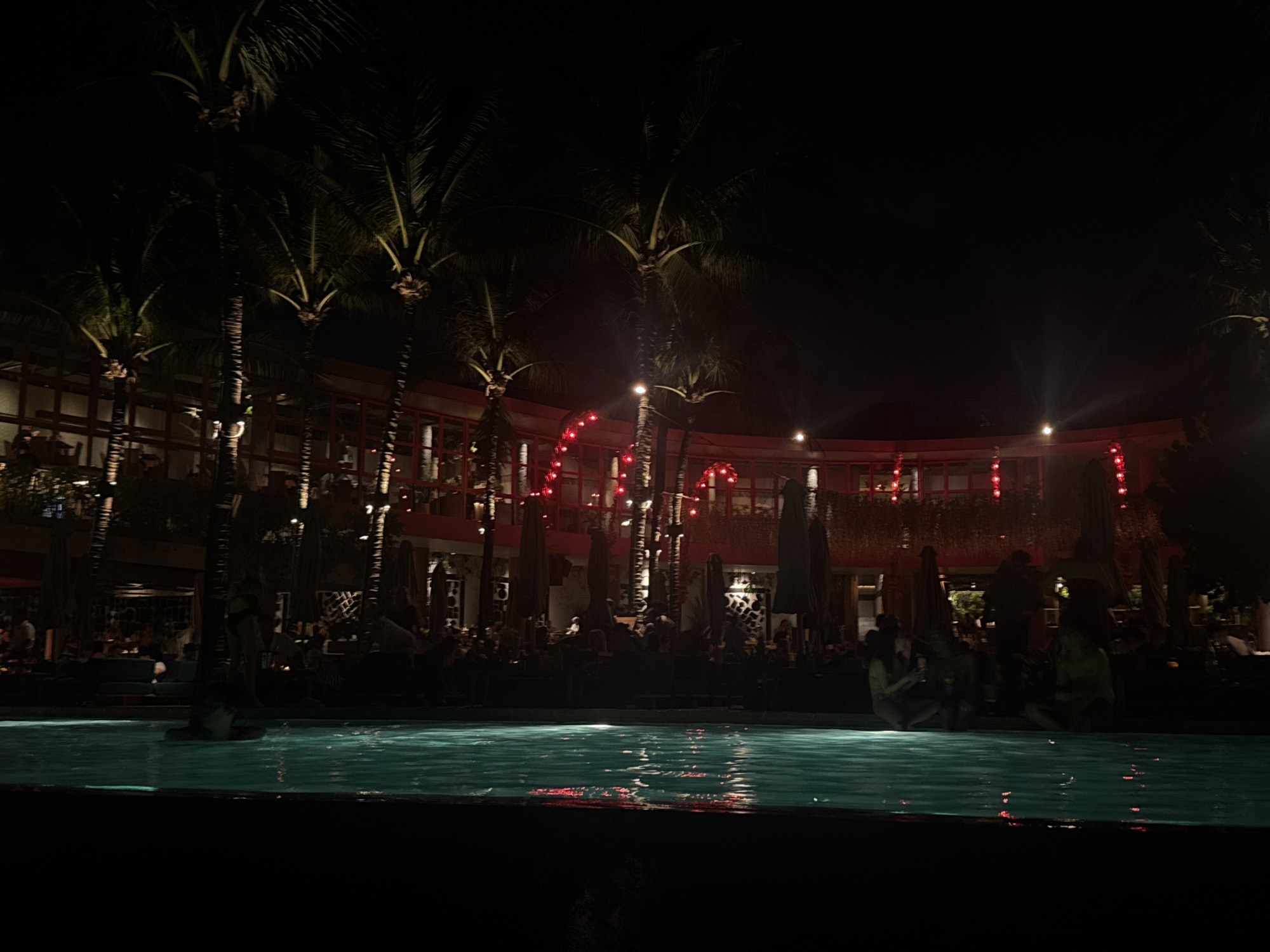The image depicts a lively night scene at a tropical resort or club, likely in North Kuta, Indonesia. In the foreground, a tranquil swimming pool with teal-colored water reflects the surrounding lights. Several individuals are visible around the pool's edge, some possibly with their feet in the water, while others are seated or standing in a brightly lit outdoor lounge or dining area just behind it.
The setting is framed by numerous tall palm trees, their dark silhouettes and fronds partially illuminated by spotlights, contributing to a tropical ambiance. Behind the lounge area, a distinctive, multi-story building with a curved, modern architectural design forms the backdrop. Its facade features numerous windows and open sections, and a prominent string of red decorative lights or lanterns traces its curved outline, casting a warm, festive glow. Additional bright lights are scattered throughout the venue, illuminating the various spaces.
The scene suggests an evening of leisure and social activity, with guests enjoying the facilities, dining, or relaxing by the pool. The time of day is clearly night, with a dark sky overhead. The atmosphere appears calm and inviting, characteristic of a high-end tropical destination. No specific text is visible in the image.