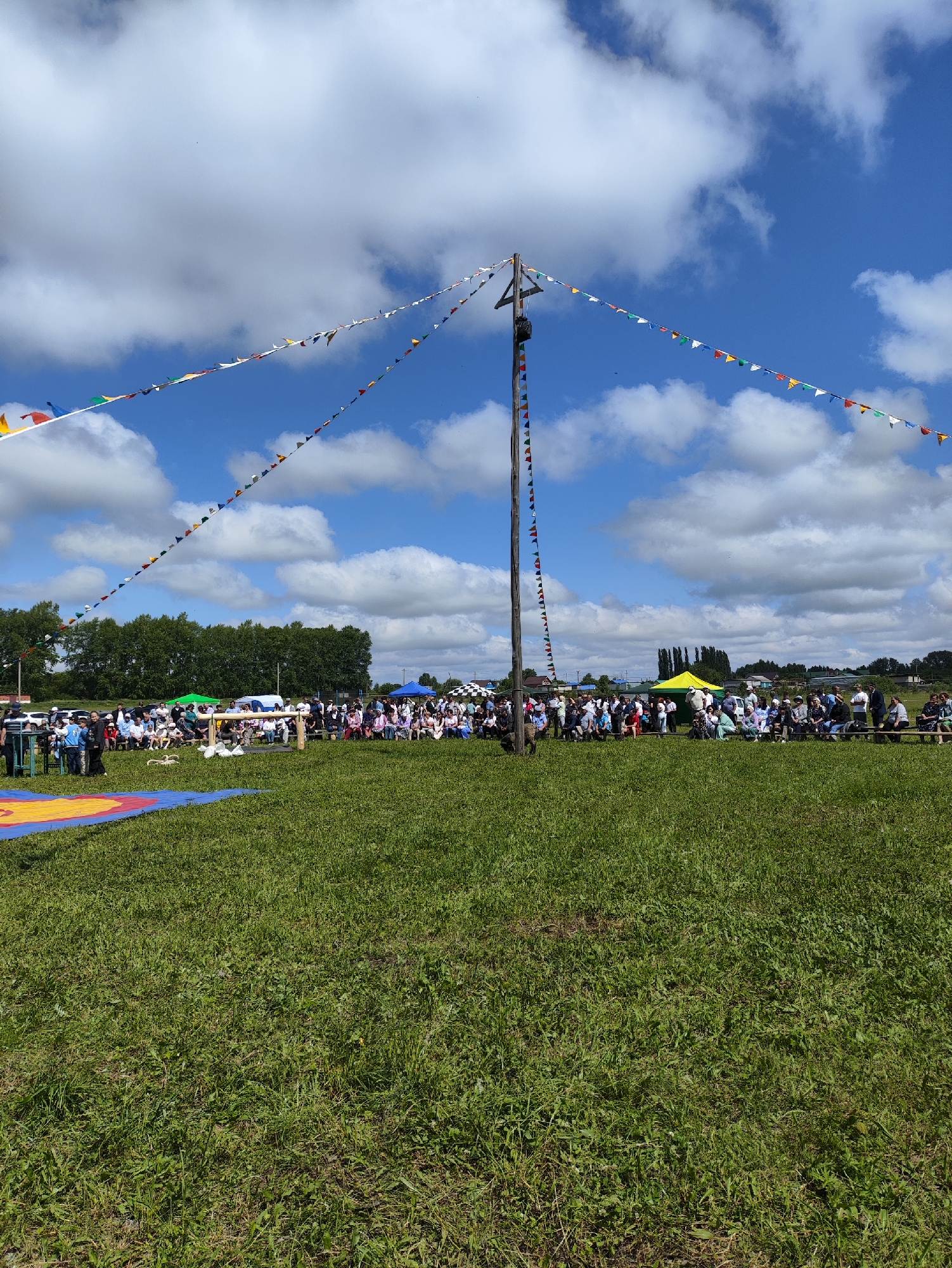 An outdoor event is taking place in a grassy field under a bright, cloudy sky. A tall wooden pole stands prominently in the center, adorned with strings of colorful triangular flags that stretch across the sky. A crowd of people are seated on benches and standing behind a wooden barrier in the middle ground, facing a wrestling mat laid out on the grass in the foreground. Several tents and some vehicles are visible in the background, beyond the crowd. The event appears to be a cultural or sporting gathering, possibly a wrestling competition or festival, taking place in the village of Novo-Ursayevo, Russia.
