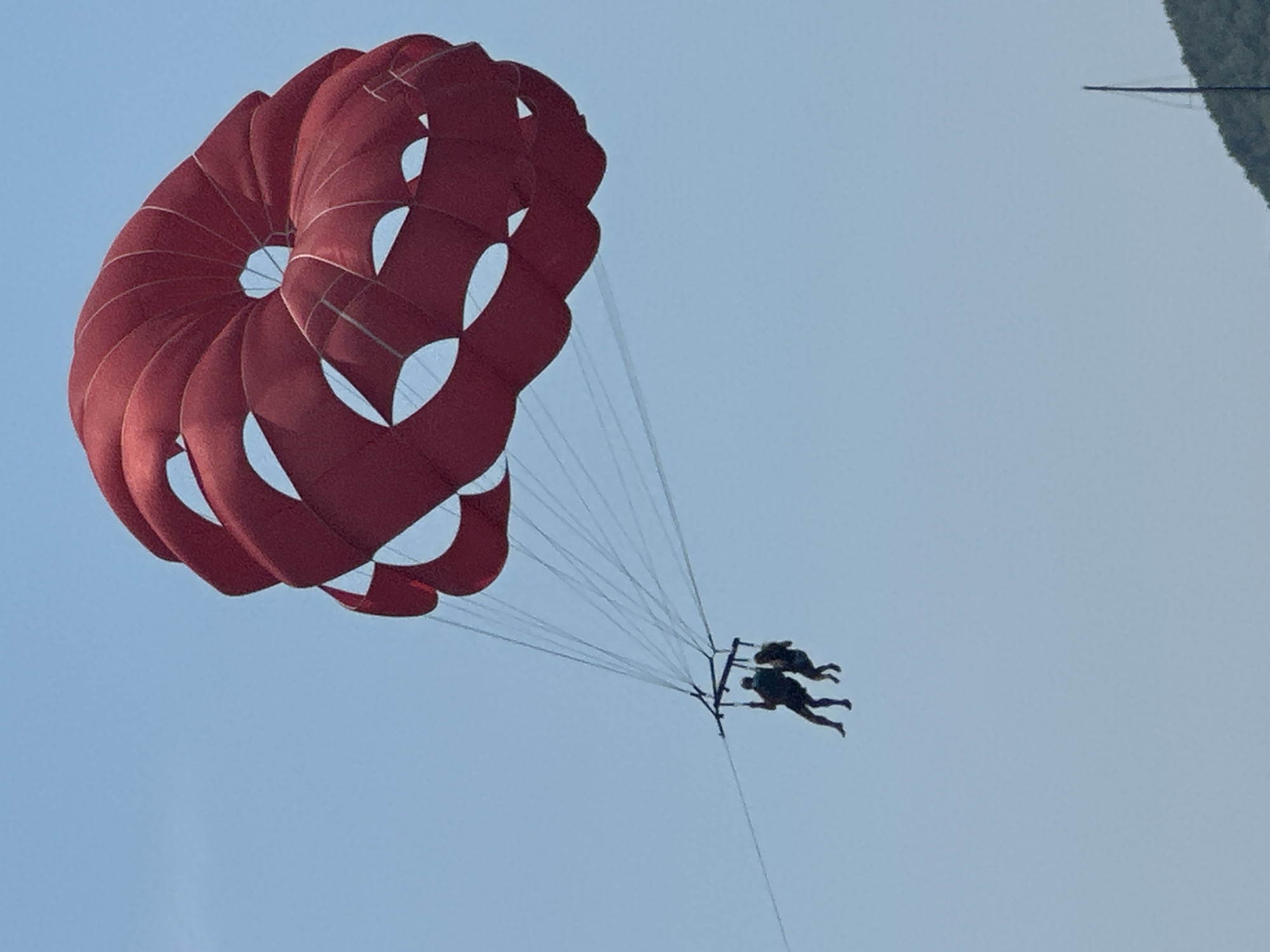 Two individuals are parasailing in the clear blue sky over Budva, Montenegro. They are suspended beneath a large, red parachute. The parachute has a distinctive circular design with openings. The two people are harnessed together and appear to be facing forward, enjoying the activity. The background is an uninterrupted expanse of sky, with a hint of a green, mountainous landscape visible at the bottom right of the frame.