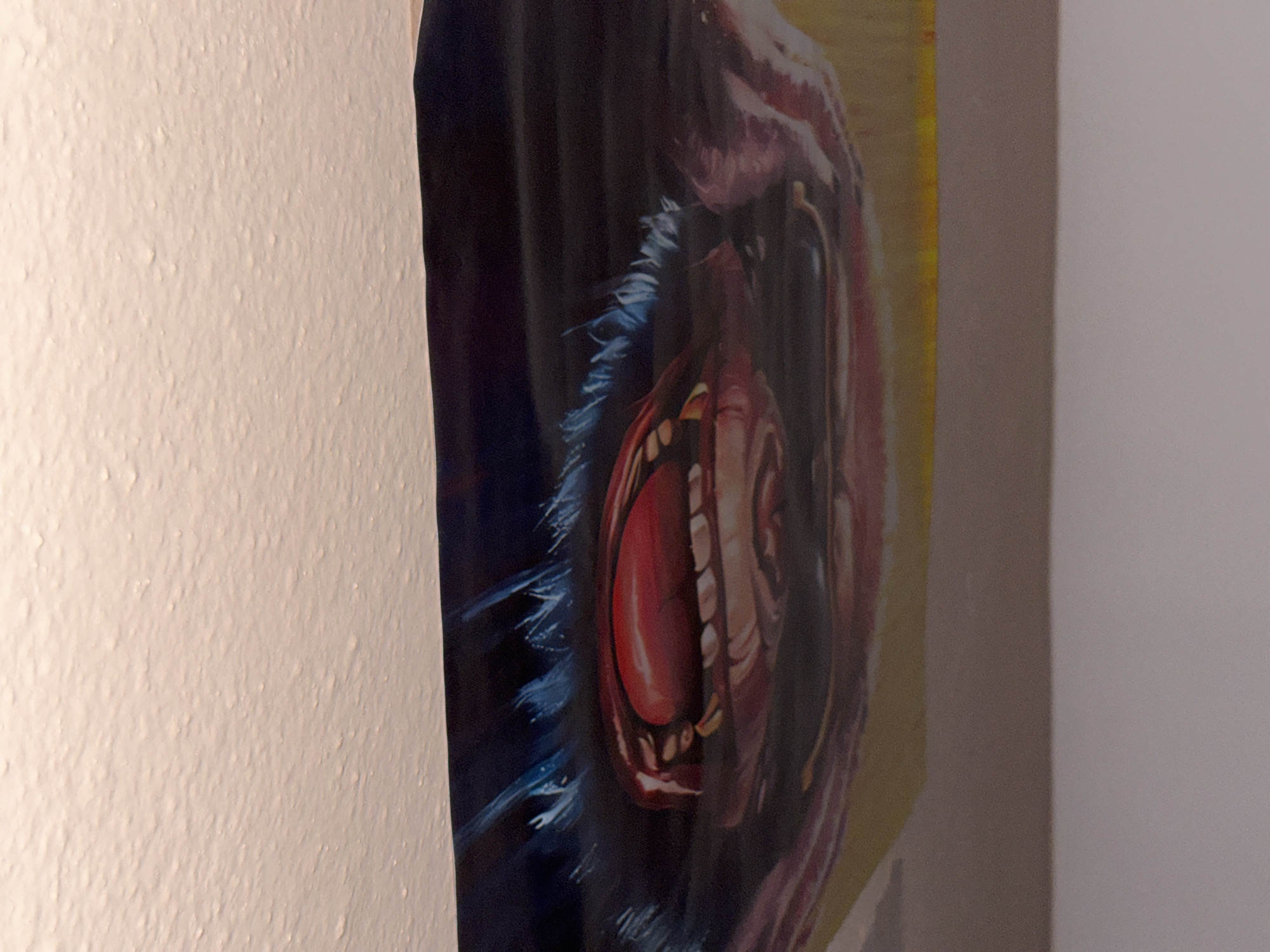 A close-up, angled photograph captures a vibrant artwork, likely a painting or poster, depicting a creature's intensely wide-open mouth. The mouth is the central and most prominent feature, revealing a large, bright red tongue, numerous sharp, pointed teeth, and a dark, cavernous interior. The area immediately surrounding the mouth is rendered in deep blue and black tones, with visible texture that suggests fur or coarse skin. To the left within the artwork, there is a darker blue-black section, while to the right, a warmer, yellowish-brown hue is visible, possibly representing the background or another part of the creature.
The artwork is mounted vertically and is photographed from a slight angle. To the immediate left of the artwork, a textured off-white wall with a stucco or popcorn finish is visible. To the right of the artwork, a smooth, plain light gray wall is seen. The lighting in the scene is soft and ambient, suggesting an indoor setting. There are no people visible, and no overt activity is taking place beyond the display of the artwork. The expression depicted in the artwork suggests a scream, roar, or intense emotion. No visible text is present in the image.