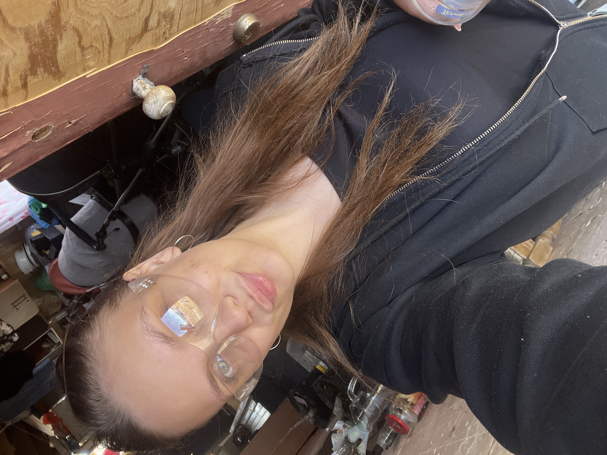 A young adult female with brown hair is wearing clear safety glasses, a black zip-up jacket, and earrings. She is holding a bottle. The background consists of a cluttered interior space with a wooden surface. The location is the city of Ольховка, Russia.