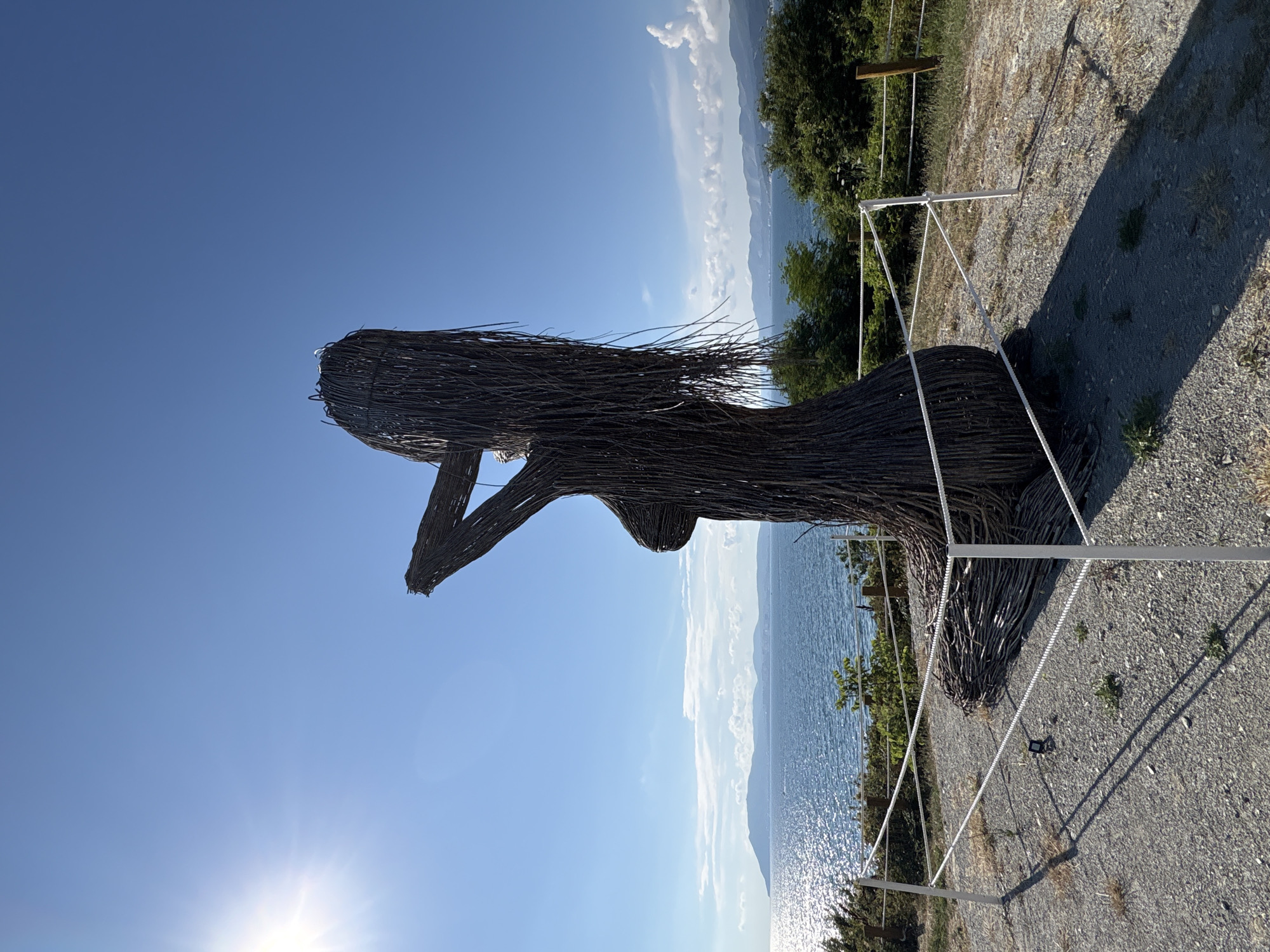An outdoor, full-shot photograph captures a large, intricately woven sculpture of a woman made from dark, natural materials, possibly branches or reeds. The sculpture is positioned on a gravelly, elevated area overlooking a vast expanse of blue sea under a clear, bright sky. The sun is shining intensely from the upper left corner, casting a bright glare and long shadows. The sculpture is partially cordoned off by white rope fencing supported by white poles, with a wooden post visible in the background. Behind the sculpture, the sparkling sea stretches towards a distant, hazy coastline with mountains. Lush green trees and vegetation are visible on the slopes leading down to the water. The overall scene evokes a sense of natural beauty and artistic expression, likely a tourist attraction in Kabardinka, Russia.