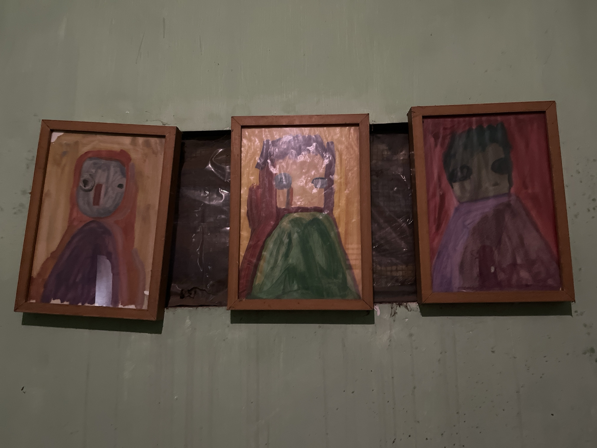 Three framed paintings of abstract figures are displayed side-by-side on a textured, light green wall in Karangkebagusan, Indonesia. The paintings, rendered in what appears to be watercolor or gouache, depict stylized human figures with simplified features. The leftmost painting shows a figure in red and purple with a grey face. The central painting features a figure with dark hair, a red torso, and a green skirt. The rightmost painting depicts a figure with dark hair, a dark face, and a purple garment against a pink background. The frames are made of light brown wood. Behind the paintings, there is a dark, crinkled material, possibly plastic sheeting. The wall shows signs of wear and has some small dark specks.