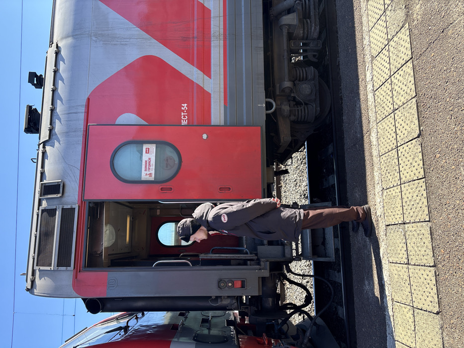 The image displays a stationary railway scene at a train station in Kropachyovo, Russia, under a clear blue sky. A male individual, appearing to be a railway employee, stands on a yellow tactile-tiled platform. He is attired in a dark brown winter coat featuring the Russian Railways (RZD) logo on the left chest, dark trousers, and dark shoes, topped with a dark winter hat. His hands are clasped behind his back, and he is oriented facing left, parallel to the train.
Immediately adjacent to the individual is a red and grey RZD passenger train car. Its main red passenger door, containing a rounded window, is open, revealing the interior vestibule. A sign visible through the window reads "ПОМНИМ ГОРДИМСЯ!" (We remember! We are proud!) accompanied by the RZD logo. The side of the train car features the RZD logo and the text "IECT-54". Another train car, primarily red and dark grey, is partially visible on the left, connected to the described car. Beneath the train car, railway tracks laid on gravel ballast are discernible. The platform transitions from yellow tactile tiles to a darker, solid surface. The environment is illuminated by direct sunlight, producing clear shadows.