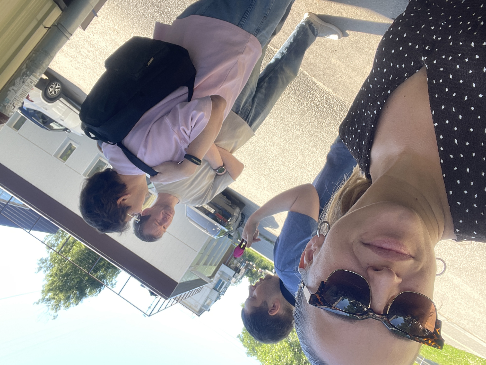 The image shows a group of four people outdoors on a sunny day, likely in the city of Chaikovsky, Russia. In the foreground, a woman with blonde hair and sunglasses is taking a selfie. She is wearing a black dress with white polka dots. Behind her, three men are visible. One man in a blue polo shirt is speaking animatedly, pointing with his left hand. Another man in a light-colored t-shirt is looking down, and a third man wearing a pink t-shirt and a black backpack is seen from the side, looking forward. The background includes a building with a dark brown awning, a white car parked on a paved surface, and some greenery. The overall setting appears to be an urban street or sidewalk.