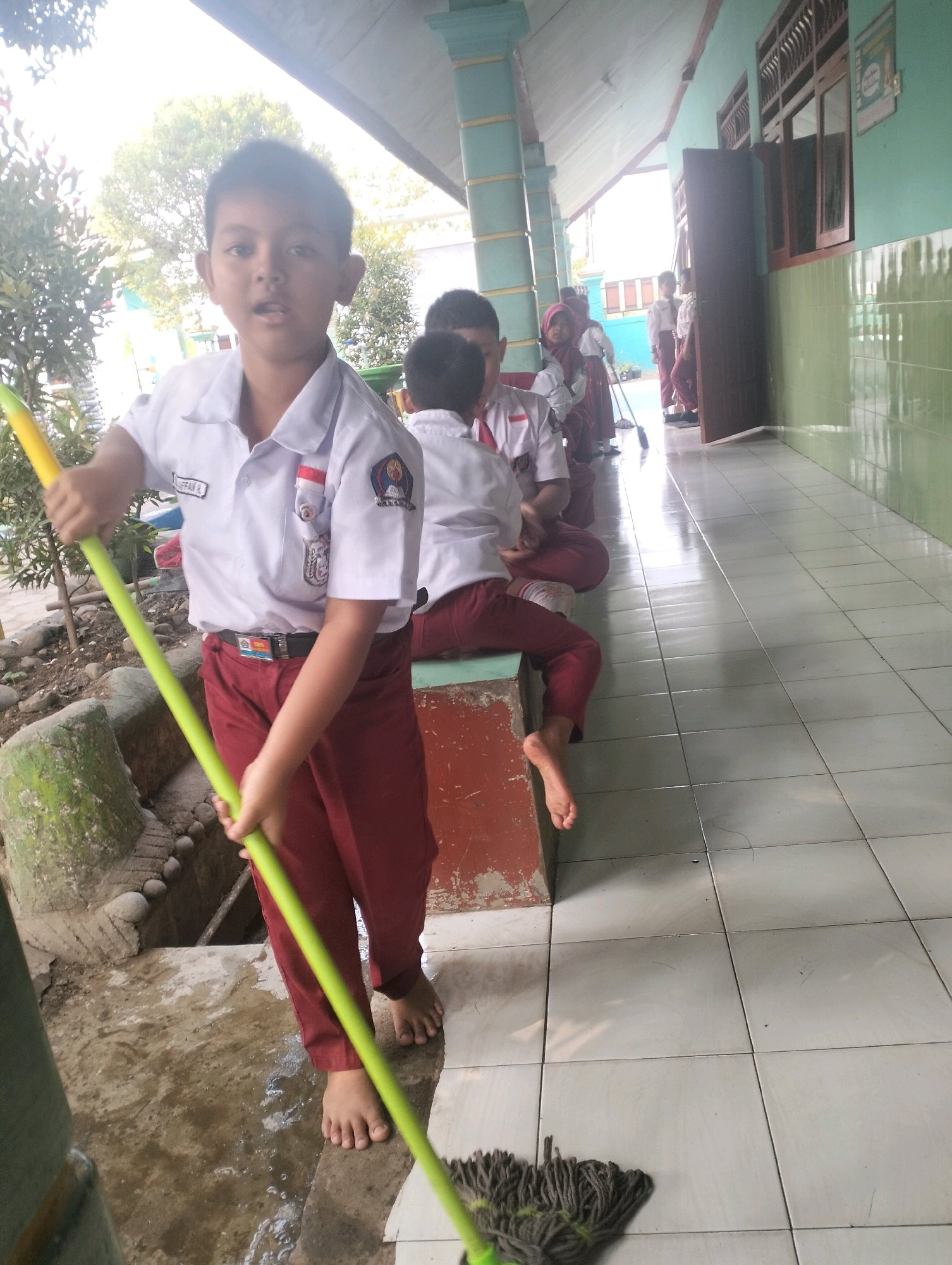 A young Indonesian boy, wearing a white school shirt with a red collar and a red sarong, stands in the foreground holding a mop with a green handle and grey mop head. He is barefoot and appears to be cleaning the ground in front of him, which is wet and dirty.
Behind him, several other children in similar school uniforms are also present. One boy in a white shirt and red pants is sitting on a concrete ledge, facing away from the camera and also barefoot. Another child, wearing a red hijab and a white shirt, is seen in the middle ground. Other children are visible further down the hallway, some standing and some sitting.
The scene appears to be a school hallway or an outdoor area adjacent to a school building. The walls are painted green and white, and there are windows and a door visible. The floor is tiled in the hallway. Sunlight is casting shadows, suggesting it is daytime. The overall atmosphere seems to be that of a school cleanup activity.
Visible text includes a name tag on the boy's shirt, "MUGIAN FA B", and a school emblem on his shirt's pocket. A small poster with text is visible on the green wall.
