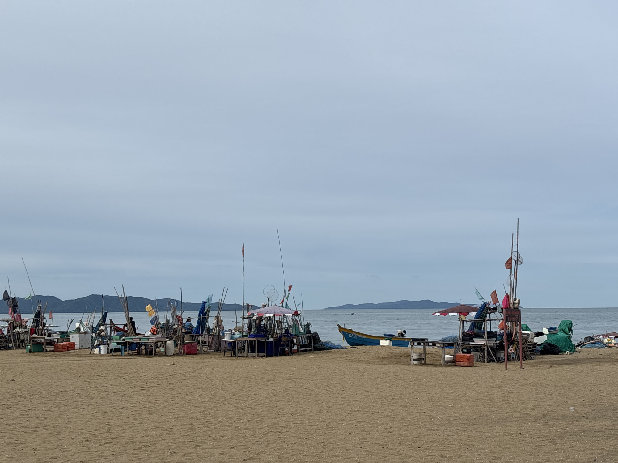The image captures a beach scene in Pattaya City, Thailand, under an overcast sky. The foreground is dominated by a sandy beach. In the midground, a collection of makeshift stalls and fishing equipment are arranged along the shoreline. These structures are built from wood and poles, adorned with various flags and umbrellas, suggesting a fishing village or market setting. Several small boats are visible, some pulled up onto the sand, while others are in the water. One prominent blue and yellow boat with an outboard motor is positioned on the sand. A few individuals are present, seemingly tending to the stalls or boats, though their actions are not clearly defined. In the background, the calm sea stretches towards a hazy coastline with hills or mountains visible on the horizon. The overall impression is one of a working fishing community engaged in daily activities.