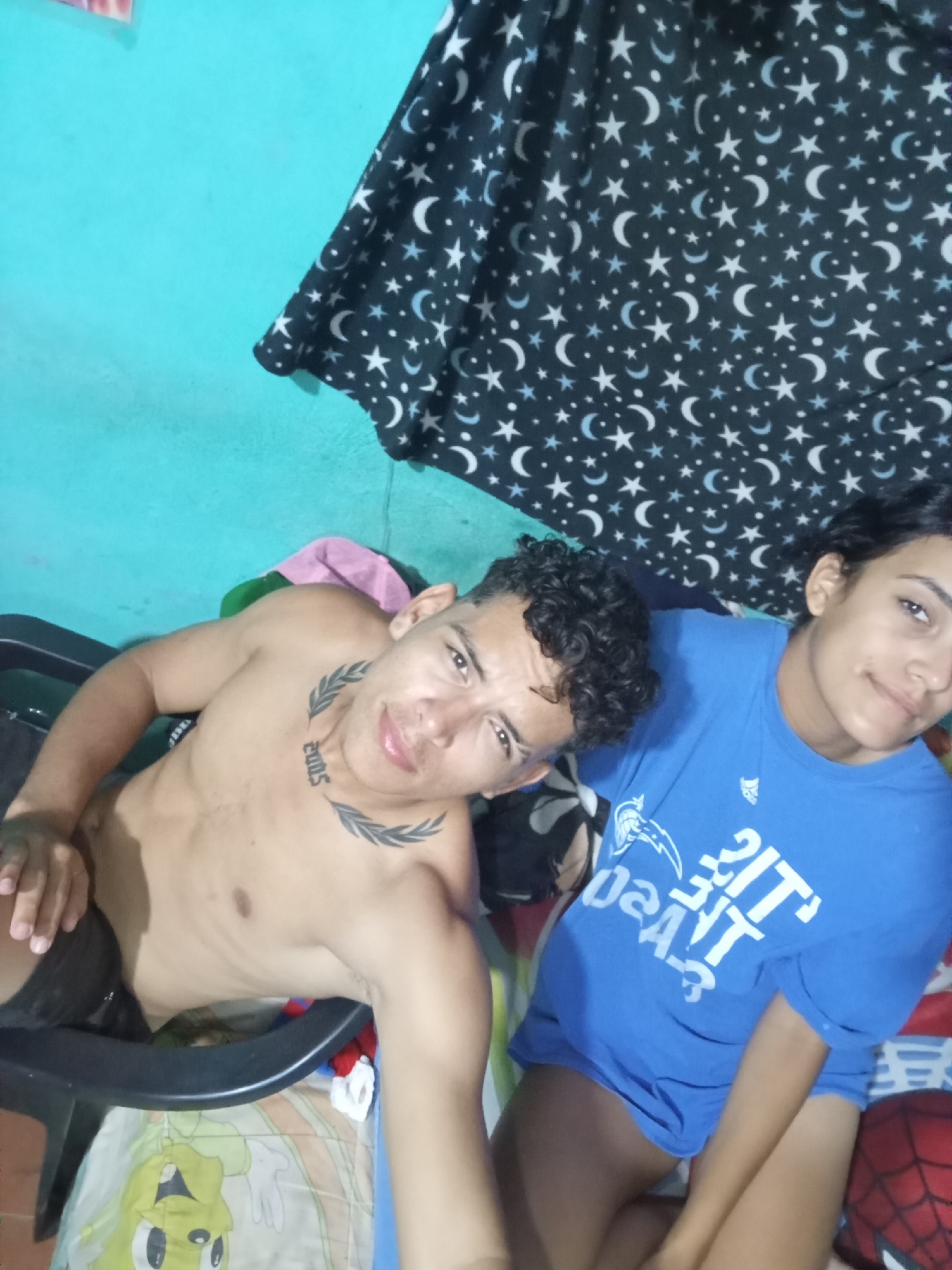 Two individuals are visible: a shirtless male with curly hair and a neck tattoo (a laurel wreath design framing the number "2005") sitting in a black chair, and a female in a blue T-shirt. The setting appears to be indoors, with a blue-painted wall and a patterned sheet with stars and moons behind them. A Spider-Man mask is visible near the bottom right of the image. The city is Altagracia, Venezuela.