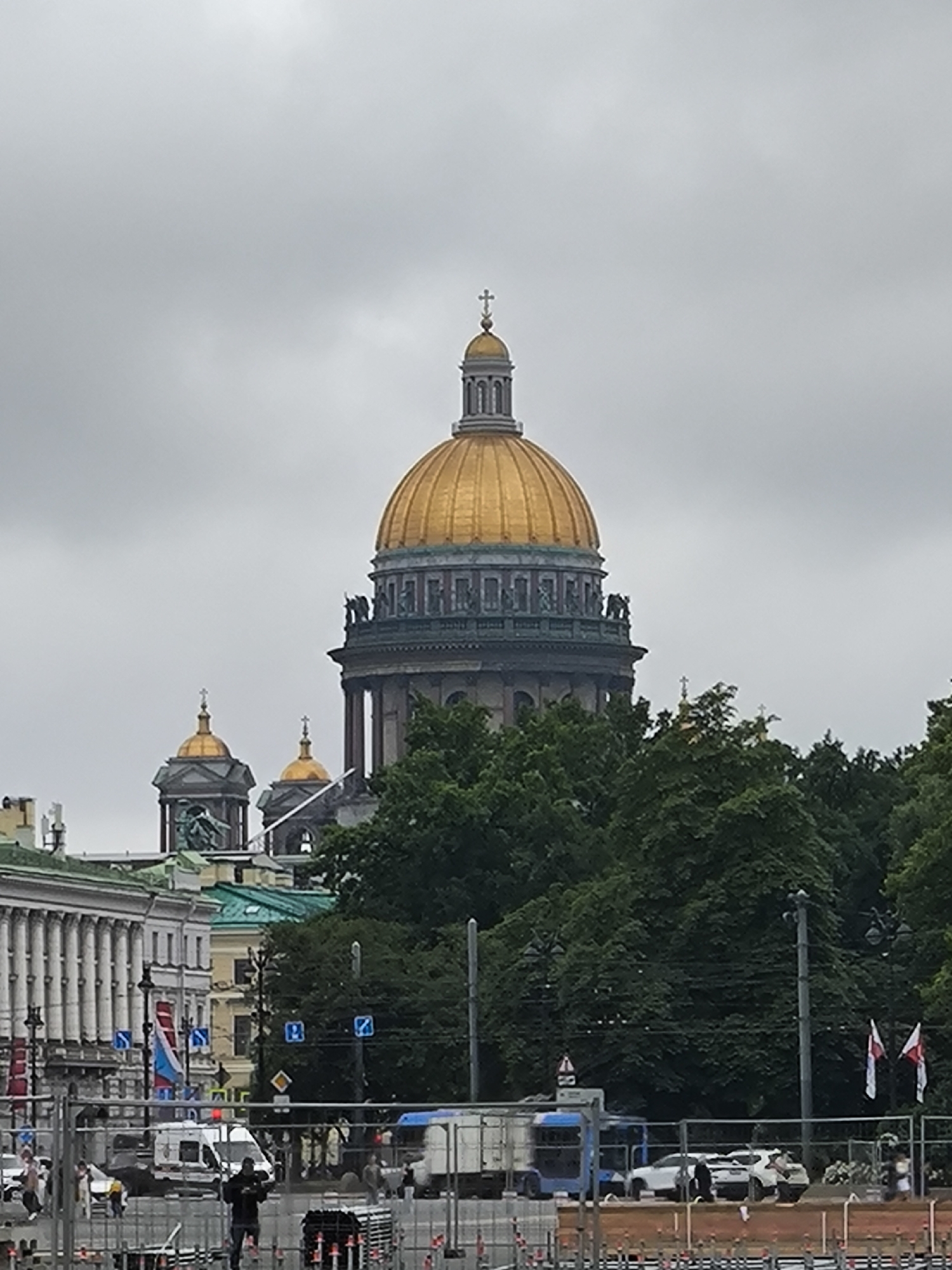 The image shows a view of St. Isaac's Cathedral in Saint Petersburg, Russia, on an overcast day. The golden dome of the cathedral is the most prominent feature, with smaller gilded domes visible to the left. In the foreground, there are trees, a street with vehicles, and construction fencing. People are visible walking and standing near the vehicles. Flags are also visible, some with red and white markings. The sky is a uniform gray, indicating cloudy weather. The architectural style of the cathedral suggests a neoclassical design.