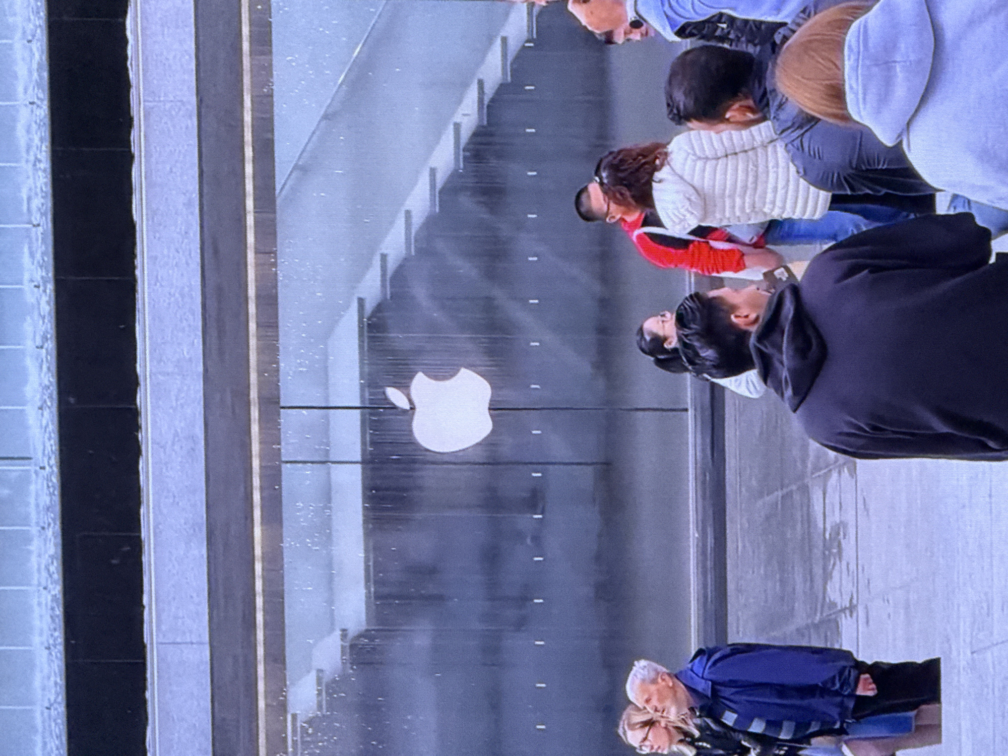 The image captures the exterior of an Apple Store, identifiable by the prominent white Apple logo on a dark glass facade. A water feature cascades down the glass, creating a shimmering effect. In front of the store, a group of individuals are gathered on a paved outdoor area. Some are looking towards the store, while others are engaged in conversation. A man in a blue jacket stands prominently on the left, while others in the foreground and midground are dressed in casual attire. The architecture suggests a modern urban setting, potentially in Dubai, United Arab Emirates, known for its contemporary design and large retail spaces.