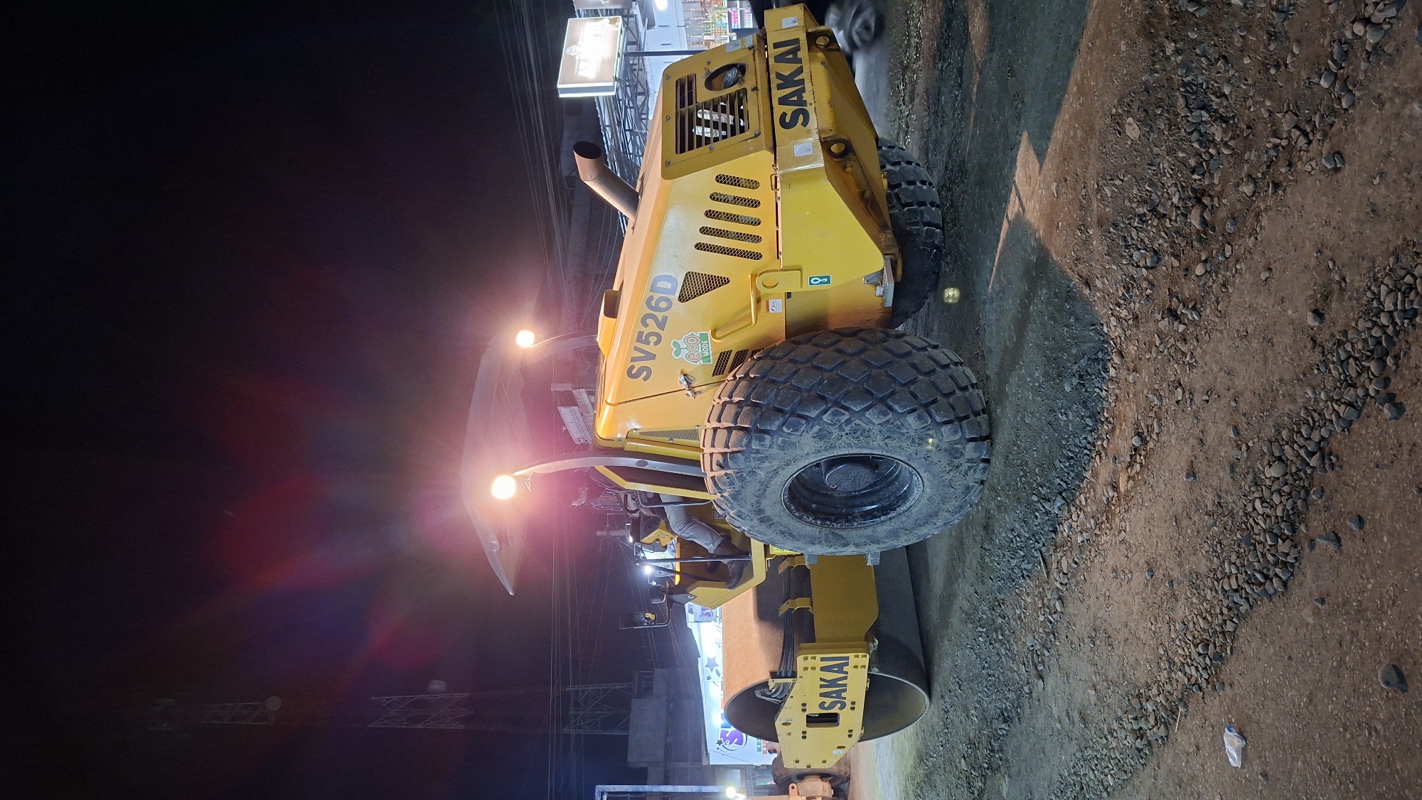 A yellow Sakai SV526D roller is parked on a construction site at night in Blangpeuria, Indonesia. The roller is the primary subject, positioned in the center of the frame, with its large, treaded tire dominating the foreground. A person is seated in the driver's seat of the roller, wearing a jacket and pants. The construction site is characterized by loose gravel and dirt, with evidence of recent work. In the background, the lights of nearby buildings and a cell tower are visible, suggesting an urban or semi-urban environment. The scene is illuminated by the roller's headlights and ambient lighting from the surroundings.