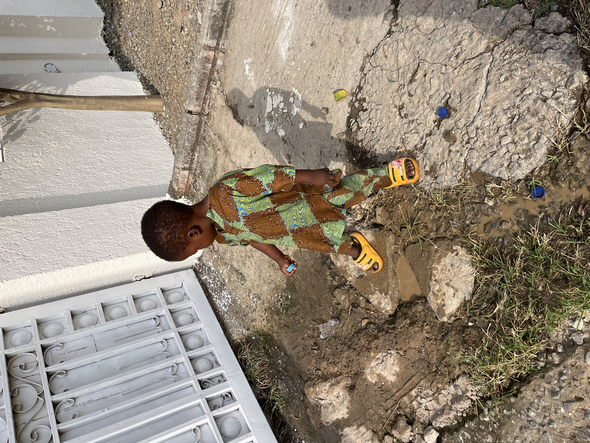 A young child in a brown and green patterned outfit and yellow sandals stands beside a gray metal gate, near a concrete path that adjoins a muddy area with scattered grass and debris. The scene is urban, potentially near residential housing in Abuja, Nigeria. The child holds a small blue object in their hand and is surrounded by bottle caps, a leaf, and miscellaneous litter. The overall setting suggests a somewhat neglected or undeveloped urban environment.