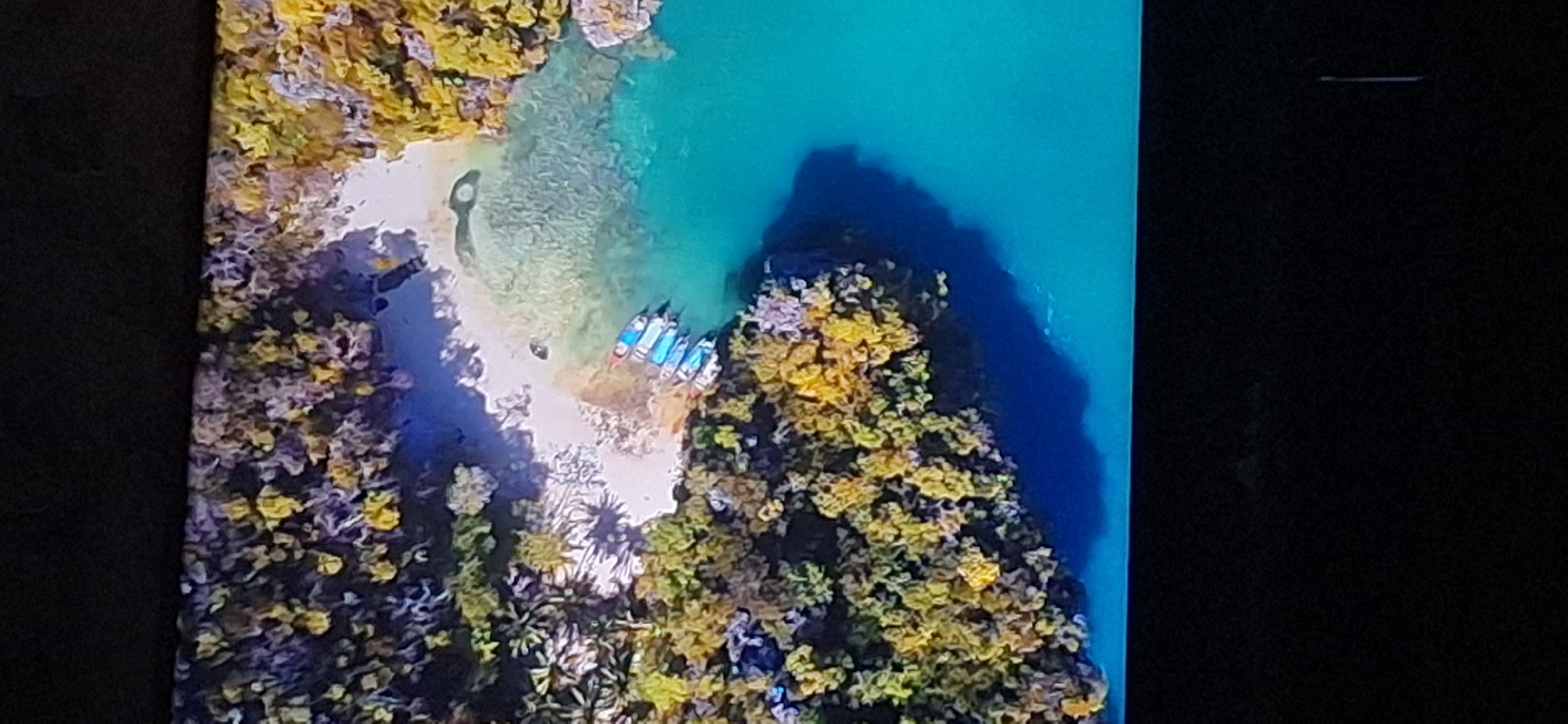 The image is a top-down aerial view of a tropical coastline with several small boats moored near a sandy beach. The water is a vibrant turquoise, transitioning to a deeper blue further from shore. Lush green and yellow foliage covers the rocky islands that surround the cove. There are no people visible in the image. This scene does not depict any location in Bila Tserkva, Ukraine.