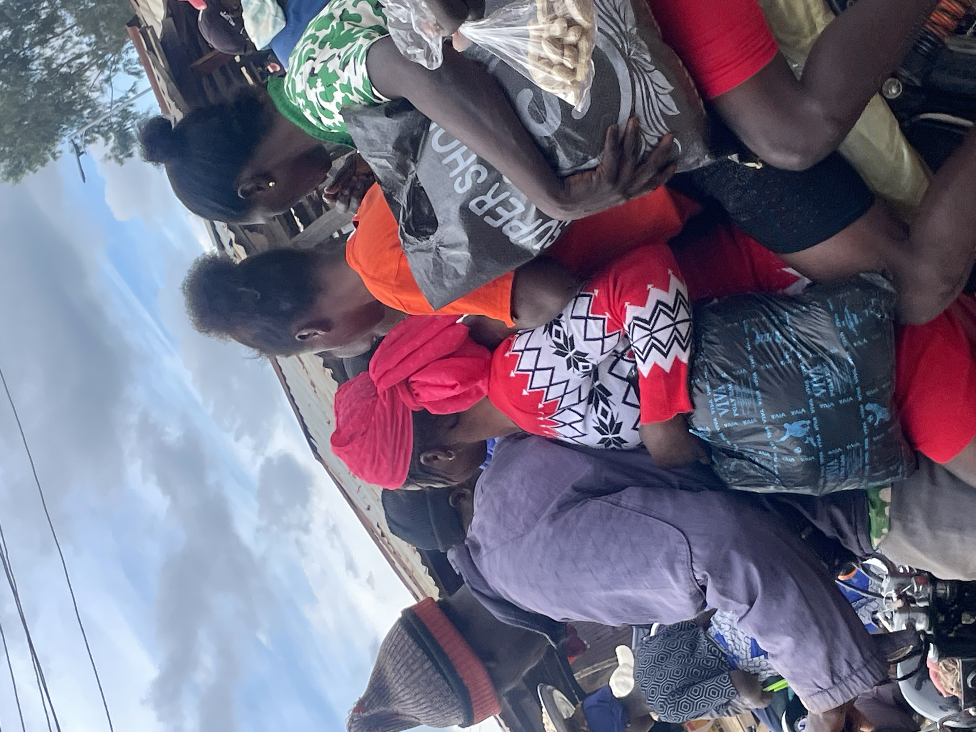 A group of people are riding on the back of a motorcycle in Wang, Nigeria, with several individuals holding bags of groceries. The motorcycle is overloaded with people and cargo, and they appear to be traveling on a dirt road. The sky is cloudy, and the weather is overcast. The buildings in the background are simple, corrugated metal structures. The people on the motorcycle are dressed in casual clothing, and they appear to be of various ages. The overall impression is one of a busy, bustling community where people rely on motorcycles for transportation.