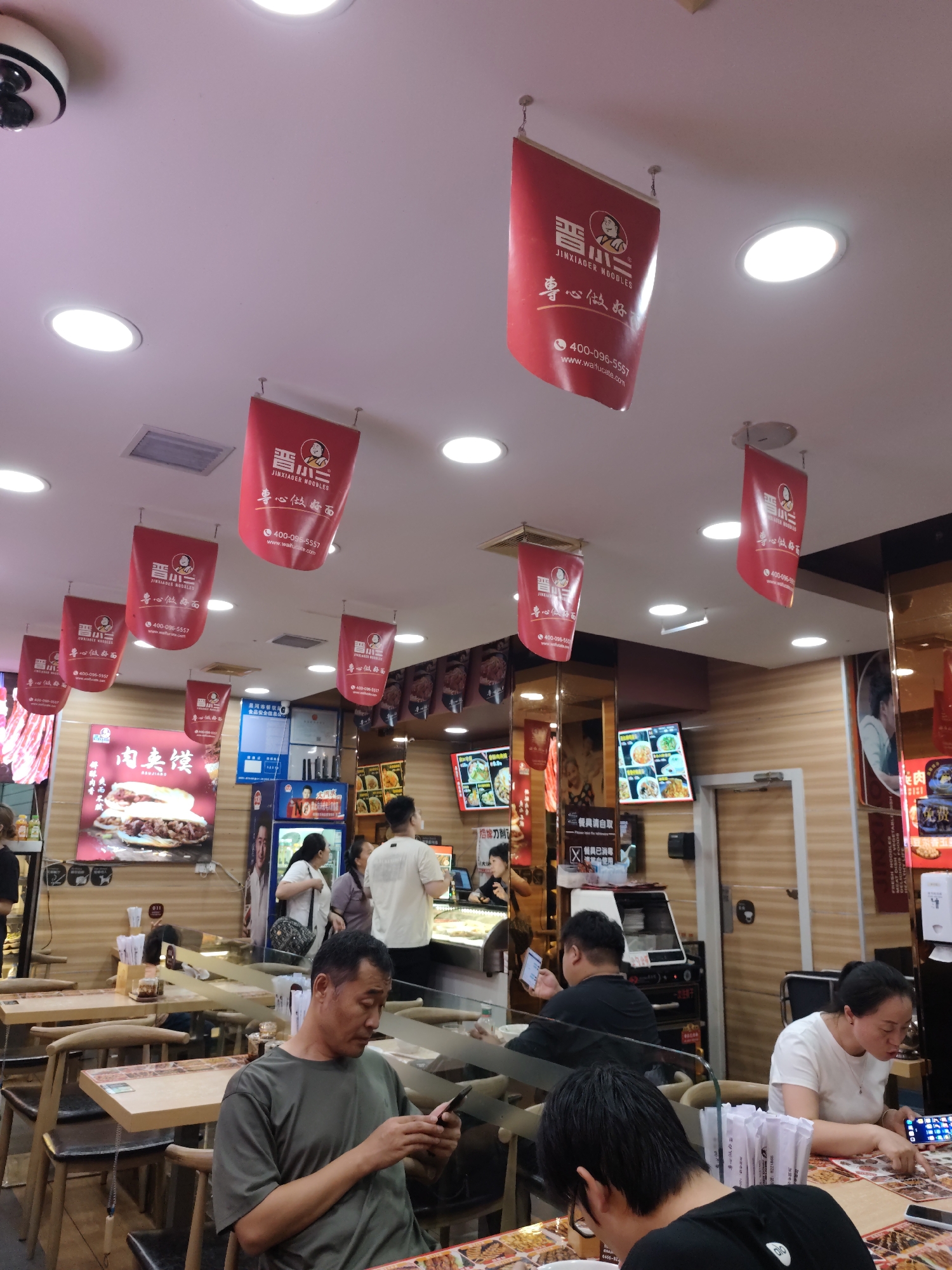 The image depicts the interior of a casual dining establishment, possibly a noodle restaurant, located in Aihui, China. The ceiling is adorned with several red banners featuring a logo with Chinese characters and "JINXIAGER NOODLES" in English, along with a phone number and website. These banners are suspended from the ceiling by wires. Round, recessed lights are visible on the ceiling, illuminating the space.
In the foreground, a man in a dark green t-shirt is seated at a table, looking at his smartphone. The table is made of light-colored wood with a patterned border and has chairs with dark seats and wooden backs. To his right, another person, a woman in a white t-shirt, is seated at a table, also looking at a device. In the background, other patrons are visible, some ordering or waiting for food, and others seated at tables.
On the wall behind the counter, there are menu boards displaying food items with images and Chinese text, including a prominent advertisement for "肉夹馍" (rou jia mo), a type of meat sandwich. A refrigerator with product advertisements is also visible. The overall environment suggests a busy, service-oriented food establishment. The wooden paneling on the walls contributes to the casual and perhaps rustic ambiance.