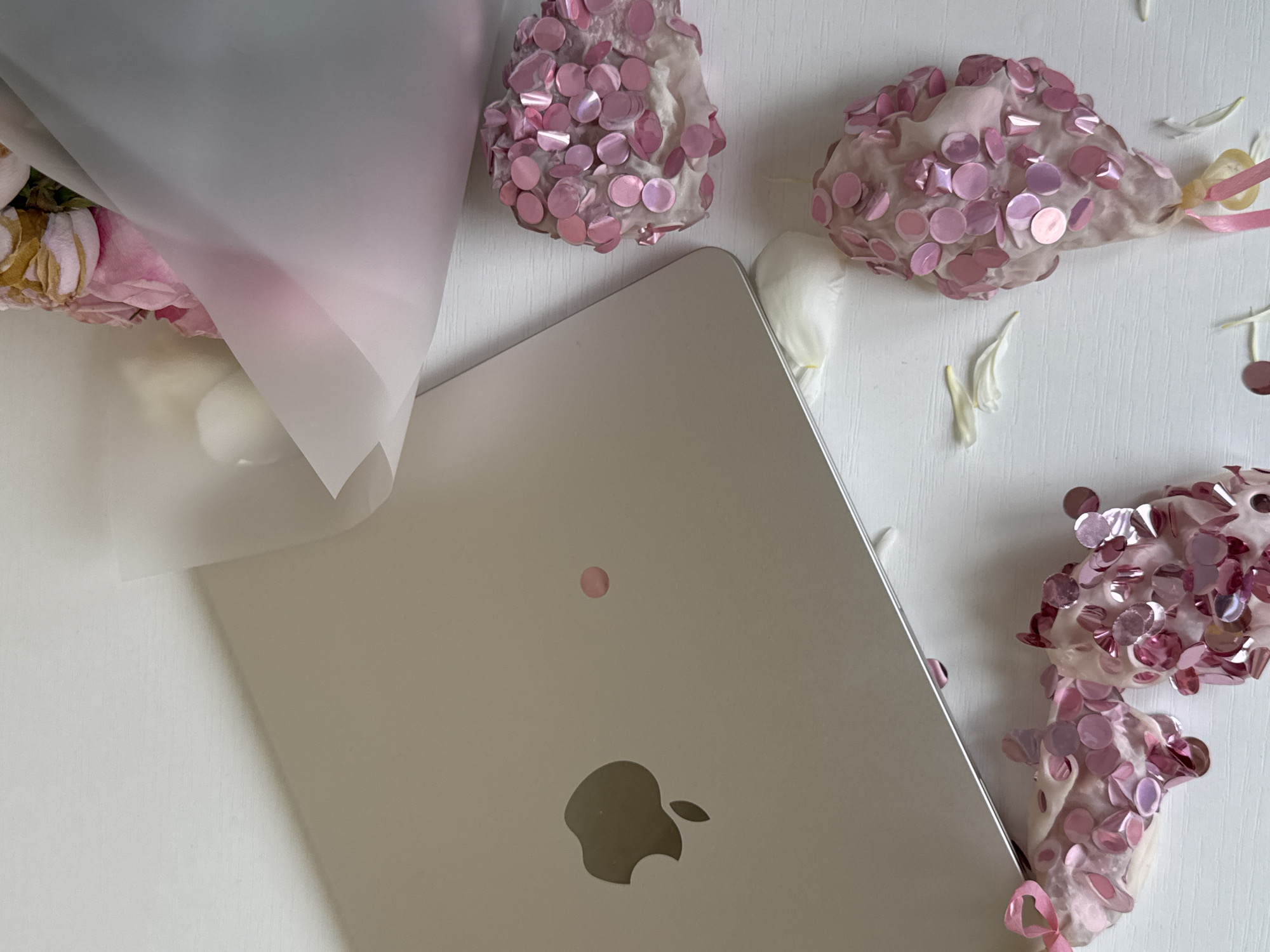 The image depicts a still life composition on a white surface, likely a table. The primary subjects are a silver laptop, a bouquet of pink and white flowers wrapped in translucent paper, and several decorative objects covered in pink sequins.
The laptop is positioned in the lower left quadrant of the image, open and viewed from above, with the Apple logo clearly visible on its lid. To the right of the laptop, towards the center and bottom of the frame, are the sequined decorative items. They appear to be some form of confectionery or craft items, shaped like clusters or small mounds, entirely covered in shimmering pink sequins of varying sizes. A thin pink ribbon is attached to one of these items.
In the upper right quadrant, a bouquet of flowers is visible, wrapped in a semi-transparent paper. The flowers appear to be predominantly pink and white, with some hints of yellow or cream. Scattered around the objects are a few white flower petals and small pink confetti circles.
The scene is lit from above, creating soft shadows. There are no people visible in the image, and no actions or interactions are depicted. The environment is clean and minimalist, emphasizing the arrangement of the objects.
The image does not provide any information that would allow for its location to be determined, including in the city of Yekaterinburg, Russia.