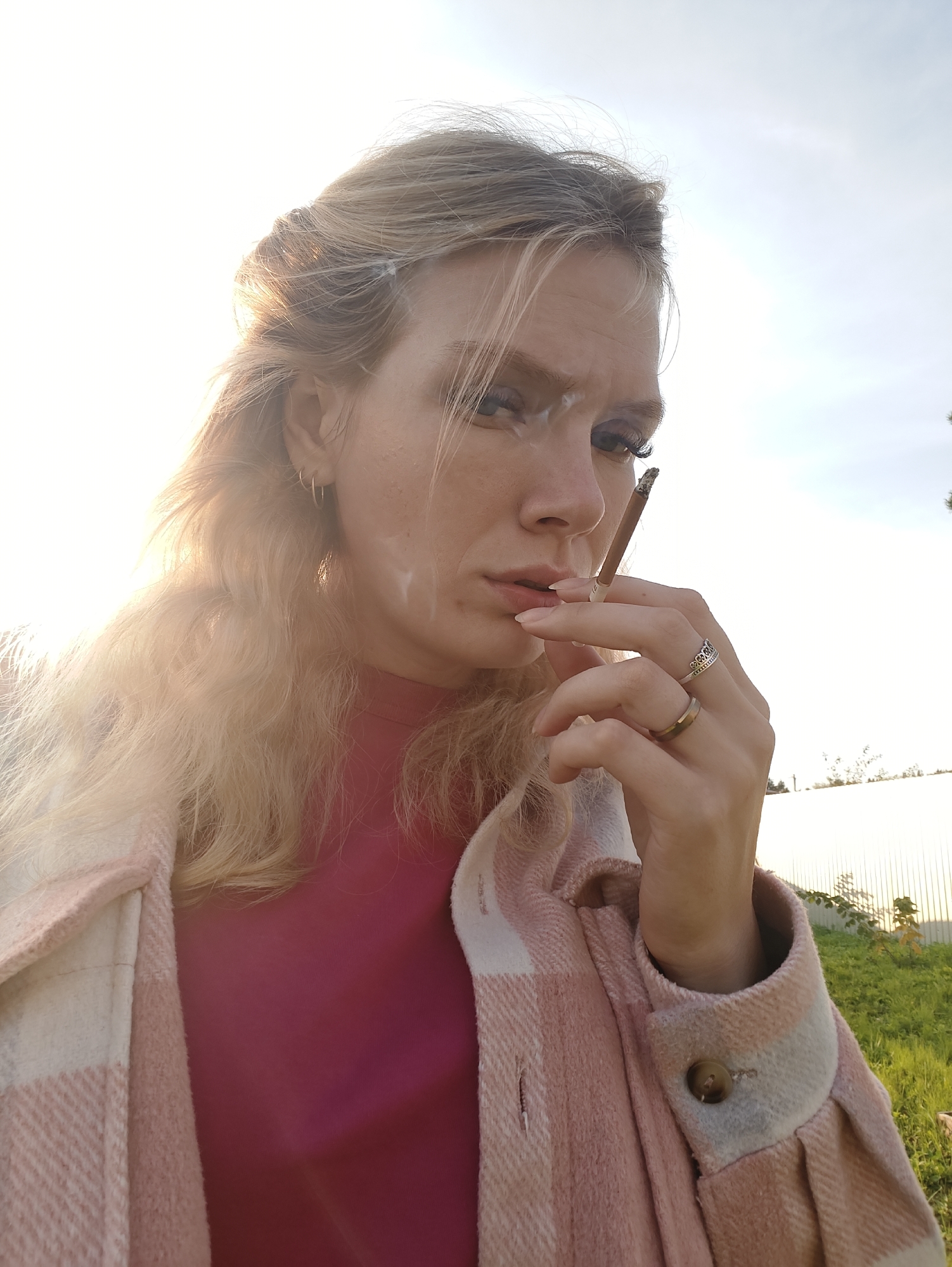 A young adult female with blonde hair is captured in a close-up, outdoor portrait. She is holding a cigarette to her lips, with smoke visible around her mouth. She is wearing a pink shirt and a pink and white checkered jacket. The background shows a bright sky, grass, and a fence. The lighting suggests it is daytime, with the sun illuminating the left side of her face.
