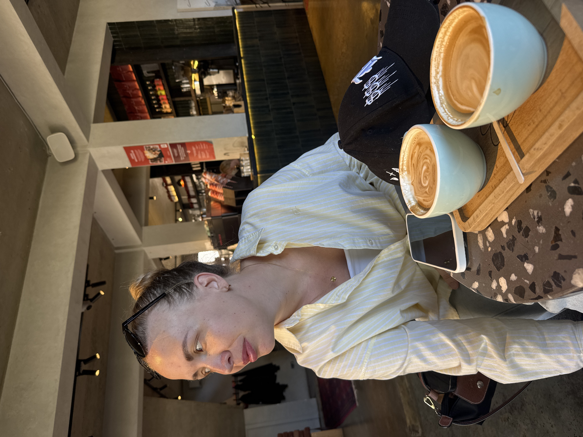 A person sits at a table in an indoor cafe in Krasnaya Polyana, Russia. The person has fair skin, sunglasses on their head, and is wearing a light yellow striped shirt and a necklace. On the table is a wooden tray with two light blue mugs of coffee, a black baseball cap, and a smartphone. The table has a patterned surface. In the background, there is shelving with bottles and other items visible, along with architectural details.
