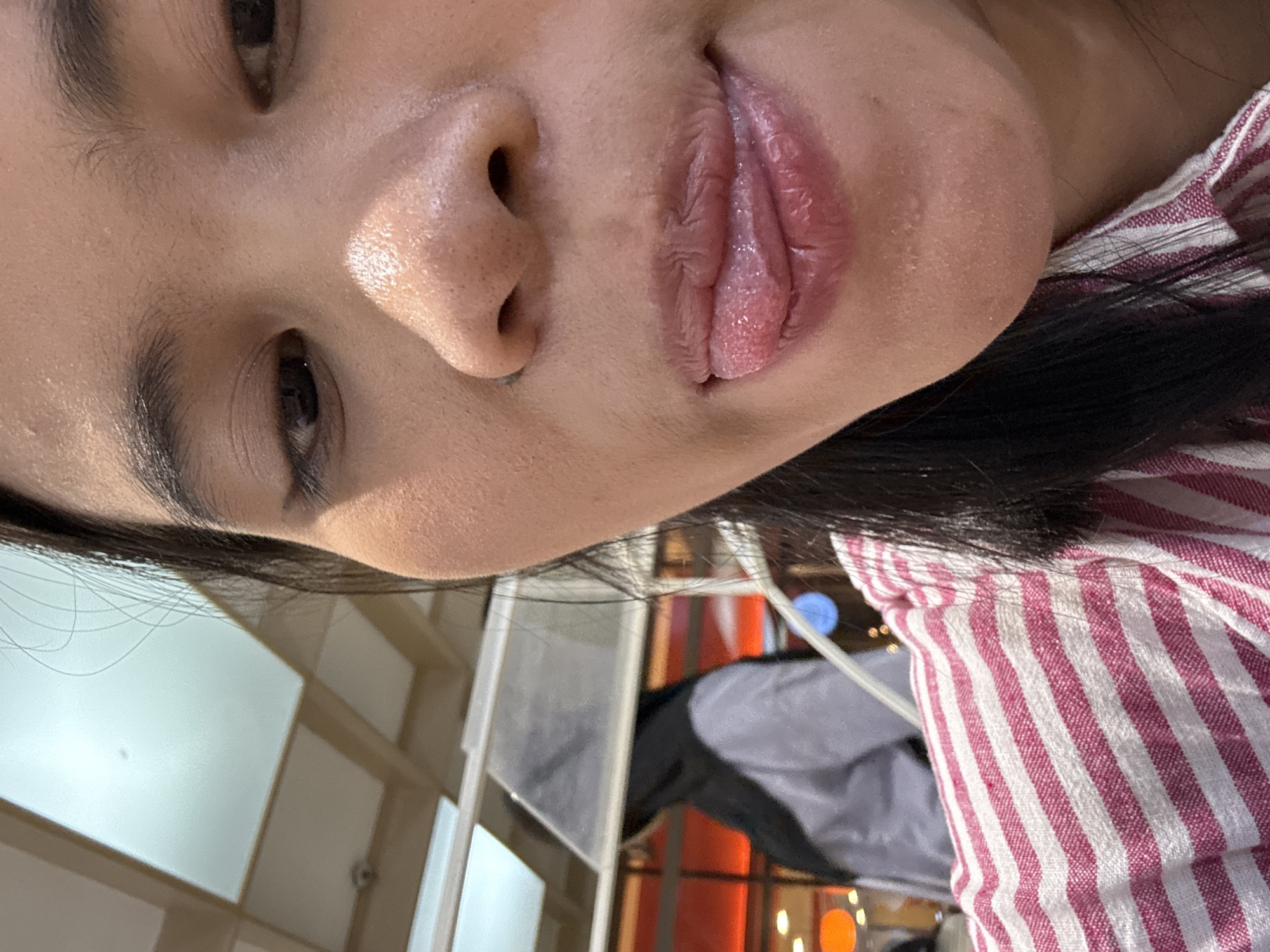 The media file is a close-up, rotated portrait of a person's face. The person's eyes, nose, and lips are visible. Their skin appears to have a light sheen, possibly from sweat or a skincare product. The person is wearing a red and white striped shirt. The background is blurred, but shows some interior elements that suggest a public space, possibly a cafe or restaurant, with some lighting fixtures visible. The lighting suggests it could be daytime or artificial lighting indoors. The overall impression is a casual, candid shot. No text is visible.