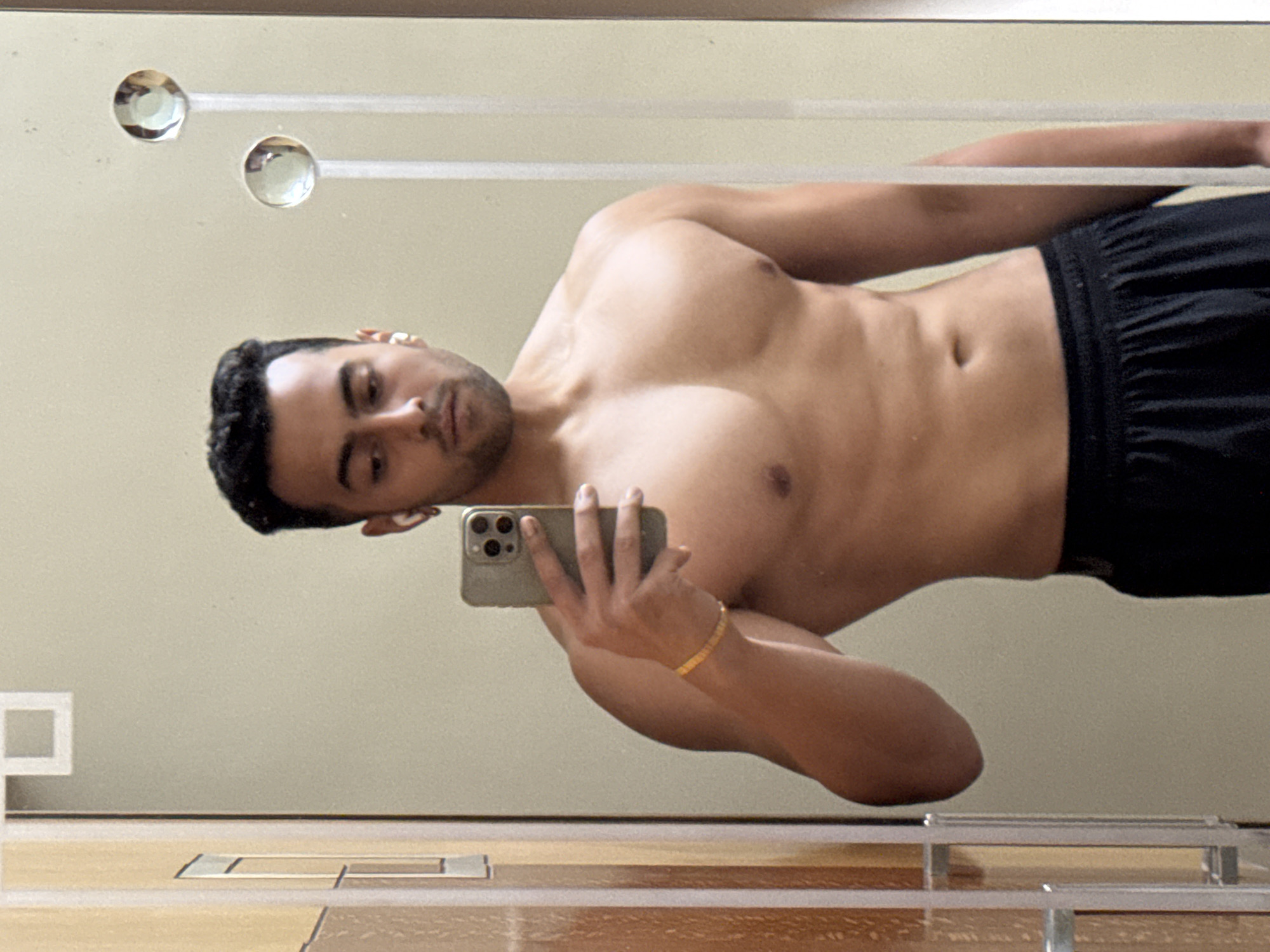 A shirtless man is taking a selfie in a mirror. He is wearing black shorts and a gold bracelet. The mirror is mounted on a wall with a neutral color, and has decorative clear knobs. The scene is possibly in a bathroom, given the vanity or shelf visible at the bottom of the mirror. The location (Kolkata, India) cannot be confirmed based on the image alone.