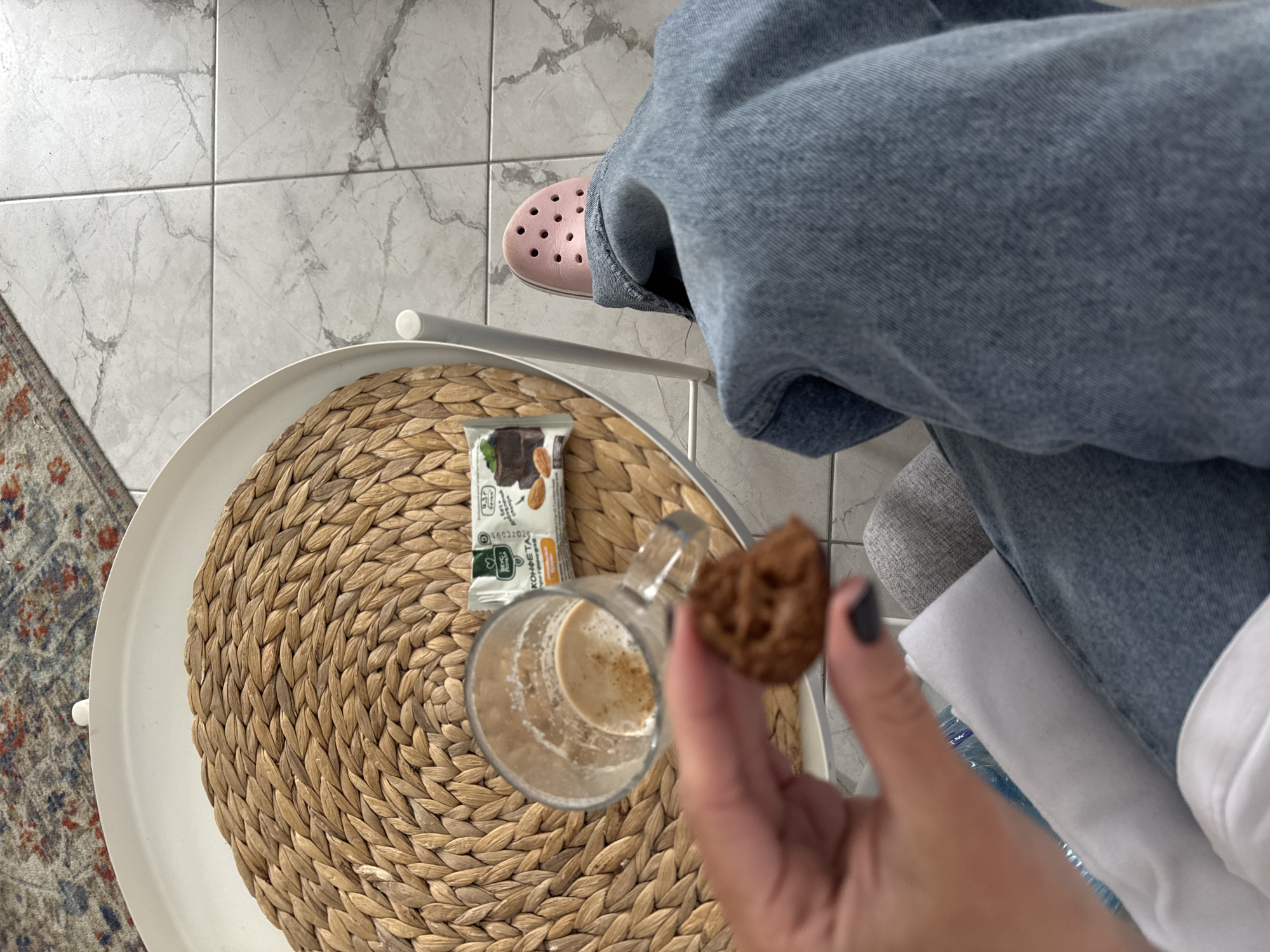 The image, set in Chaikovsky, Russia, presents an overhead view looking down at a person's lower body and a small side table in an indoor environment.
In the foreground, a left hand, with dark grey nail polish, holds a small, dark brown, irregularly shaped cookie or confection. Below the hand, the person wears faded blue denim jeans, with a grey fabric cushion or blanket visible on their lap, and a white fabric item and a partial blue plastic bottle also present. To the right, a right foot is visible, wearing a light pink Crocs-style sandal with visible ventilation holes and a white sole strip.
A round white metal side table is positioned to the left and center, featuring a circular woven placemat made of natural fibers. On the placemat rests a clear glass mug containing a light brown, frothy, partially consumed beverage, likely coffee or a latte, with a dusting of spice on top. Adjacent to the mug is a packaged snack bar, green and white, with Cyrillic text reading "ВКУС ПОЛЬЗЫ" (Taste of Benefit/Usefulness) and "КОНФЕТА" (Candy/Sweet). The packaging depicts a chocolate-like bar with almonds and indicates "4,3 г" (4.3g), likely a nutritional value.
The flooring is a grid of light grey ceramic tiles with a subtle marble-like veining pattern. In the upper left background, a portion of a patterned rug with red, blue, and beige tones is visible.