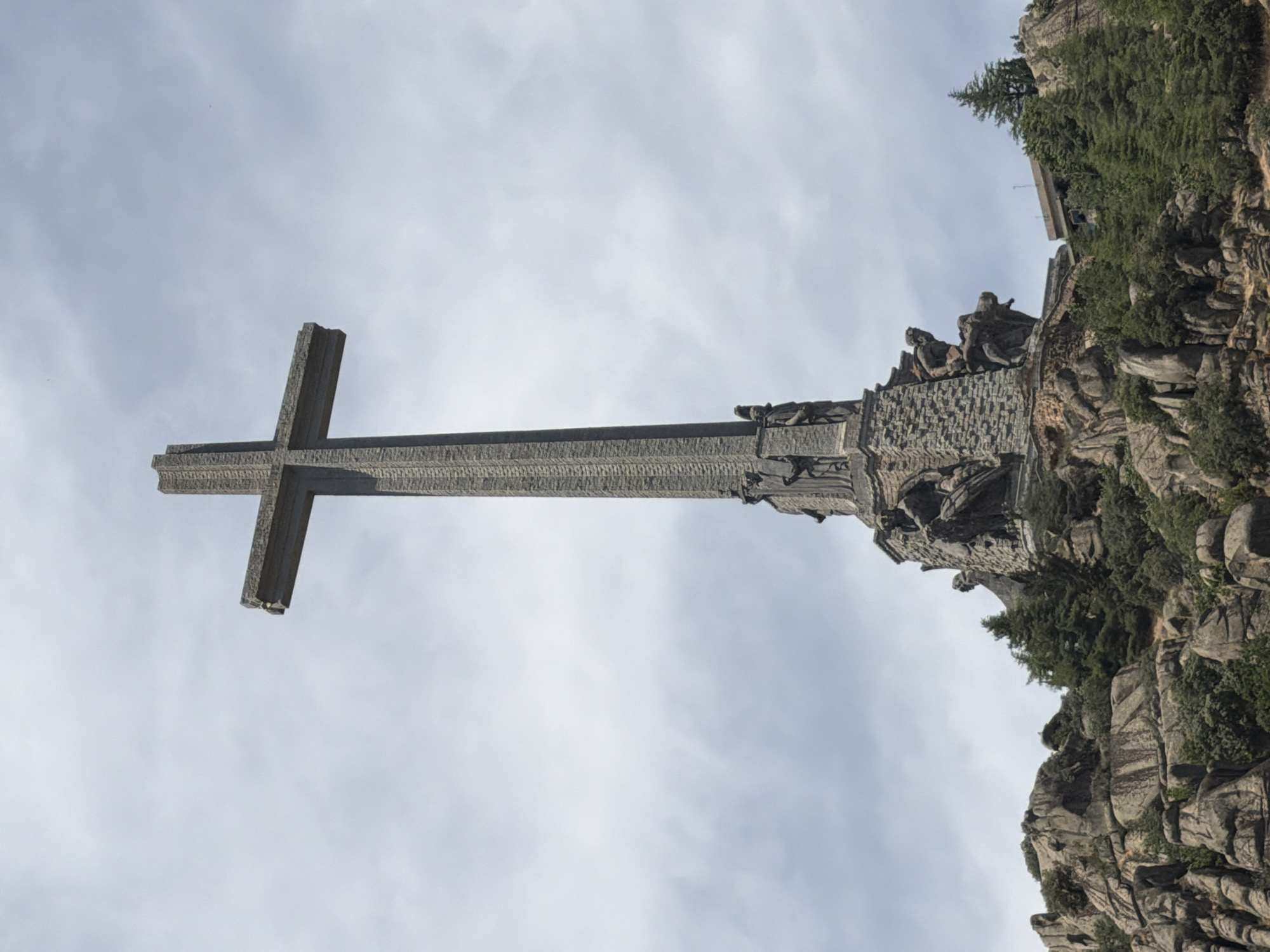 A towering concrete cross monument stands prominently on a rocky hilltop, set against a pale, cloudy sky. The monument, constructed from stone blocks, features detailed sculptures at its base, depicting figures and scenes. The cross itself is a large, simple structure with a thick central shaft and a horizontal beam. The surrounding environment is rugged and mountainous, with scattered trees and rocky outcrops. A building is visible in the background, to the right of the monument. The monument is the Valle de los Caídos (Valley of the Fallen), located in the Sierra de Guadarrama mountain range near San Lorenzo de El Escorial, Spain.