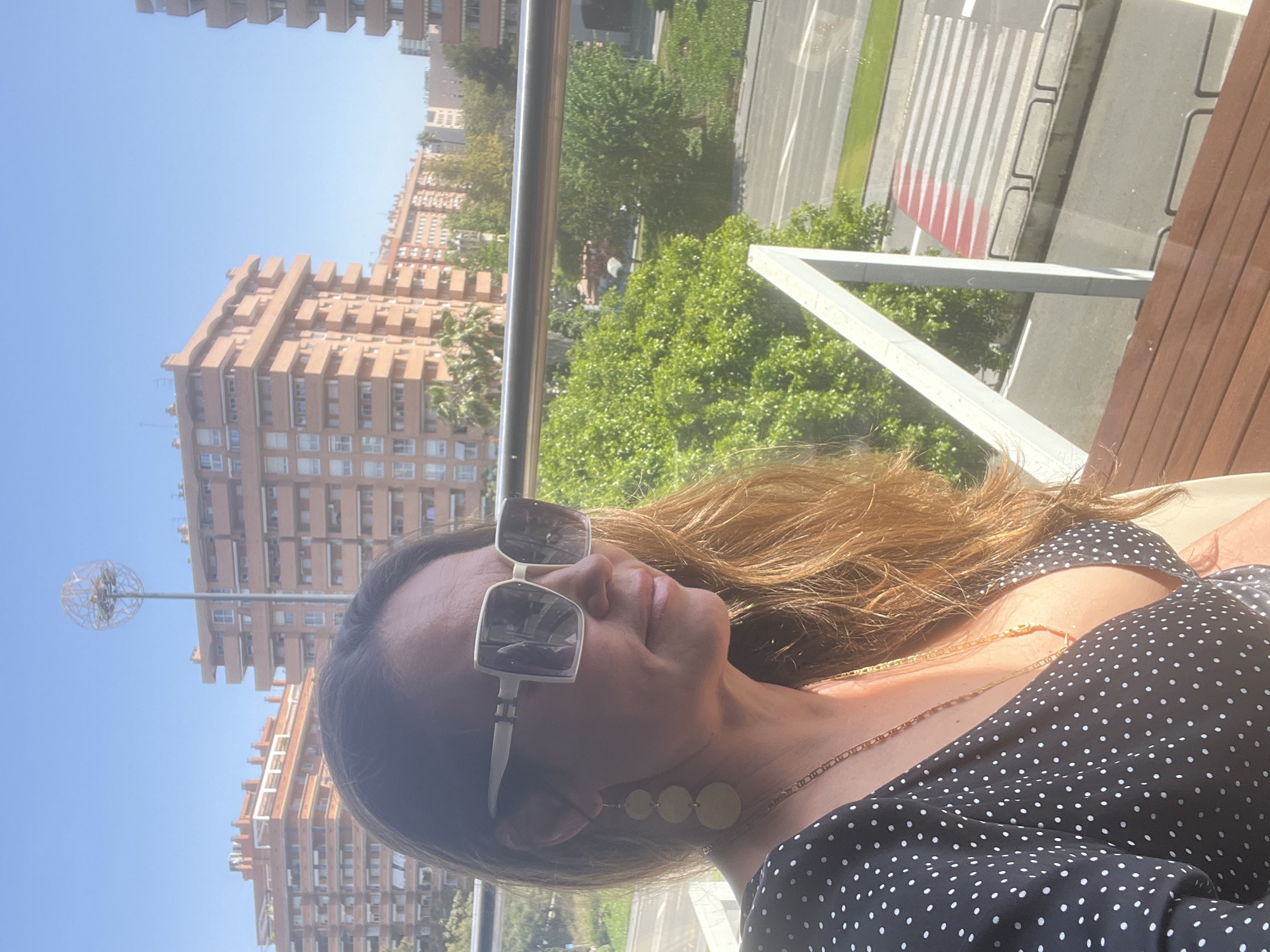A medium-range, rotated, sunlit outdoor portrait shows a woman in a black-and-white polka dot dress and large sunglasses, facing right. She wears a gold necklace and disc earrings. The background is the city of Tarragona, Spain, with tall salmon-colored apartment buildings, green trees, and a road with crosswalk. There is a metal railing and wooden tabletop in the foreground.