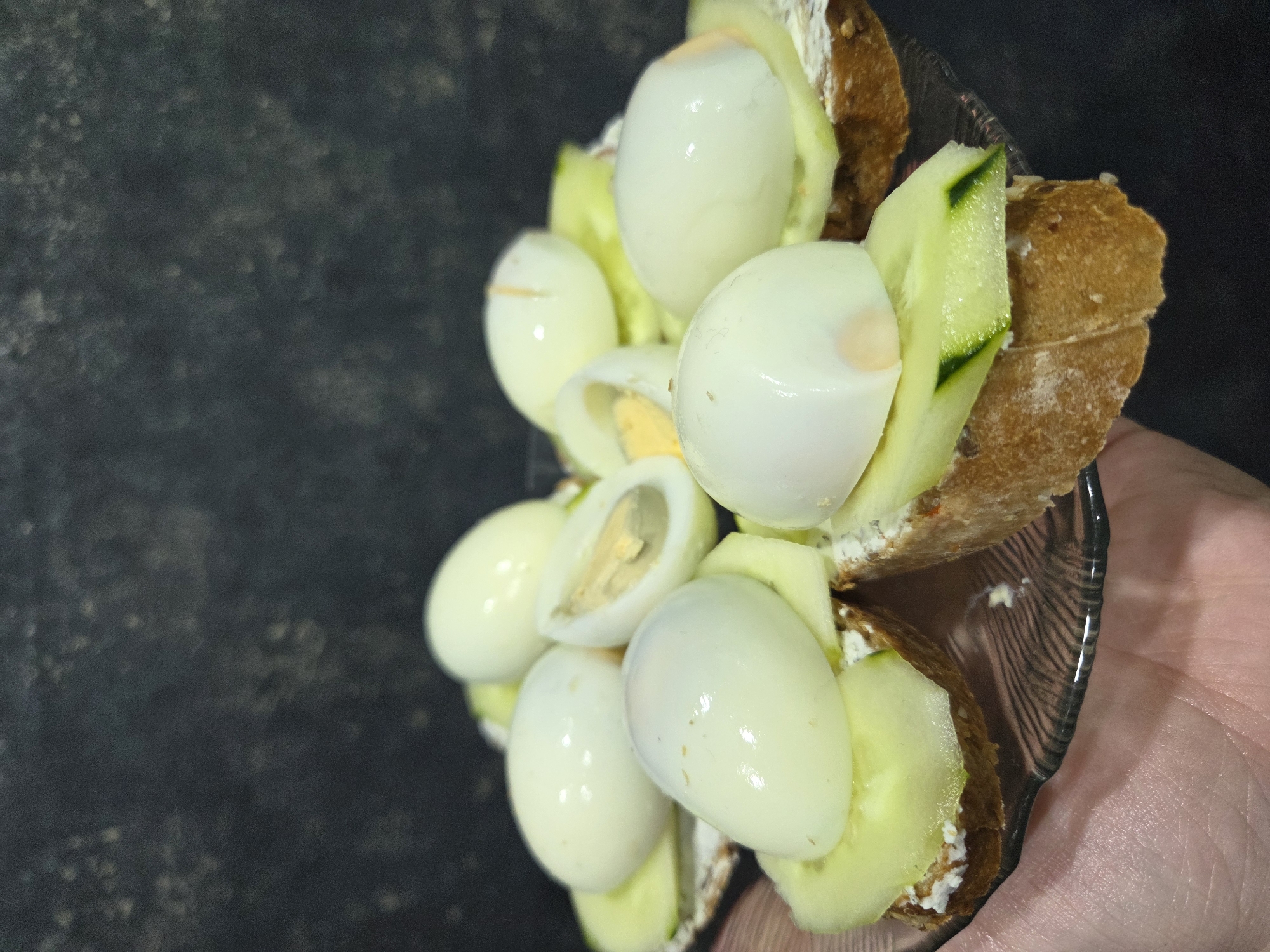 A close-up, overhead shot captures a small, clear glass plate holding several open-faced sandwiches. The sandwiches consist of slices of dark, crusty bread topped with a white spread, thin slices of green cucumber, and halves of hard-boiled quail eggs. Some of the eggs are whole halves, while one is sliced in half vertically, revealing its yellow yolk and white. The background is a dark, textured surface, likely a wall or tablecloth, with a subtle dappled pattern. A human hand, with visible skin texture and veins, is holding the edge of the plate from the lower left. The lighting is somewhat dim, creating soft shadows and highlighting the sheen on the egg whites. There is no discernible indication of the specific location within Zheleznogorsk, Russia.