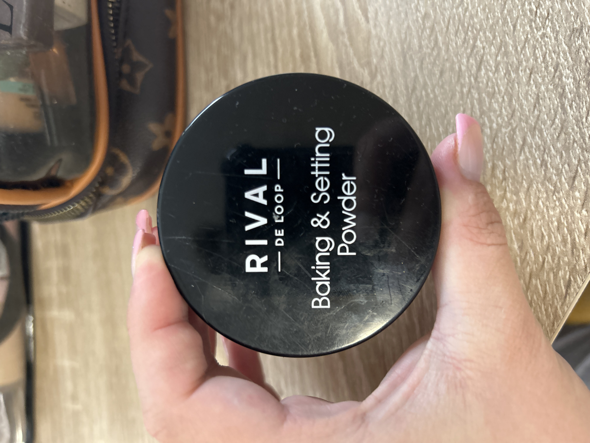 An overhead close-up image, captured in Waldbröl, Germany, features a fair-skinned hand holding a black cylindrical cosmetic container. The hand, exhibiting light pink nail polish on the visible fingernail, grips the container against a light-colored wooden or laminate surface. The container's lid is black and shows multiple surface scratches. White sans-serif text is printed on the lid: "RIVAL" at the top, followed by a horizontal line and "DE LOOP" beneath it. Below this, "Baking & Setting Powder" is displayed in two lines. In the blurred background, towards the upper right, a brown and tan patterned cosmetic bag with orange trim and a zipper is partially visible, containing indistinct objects. The environment appears to be an indoor setting, possibly a vanity or dressing table.