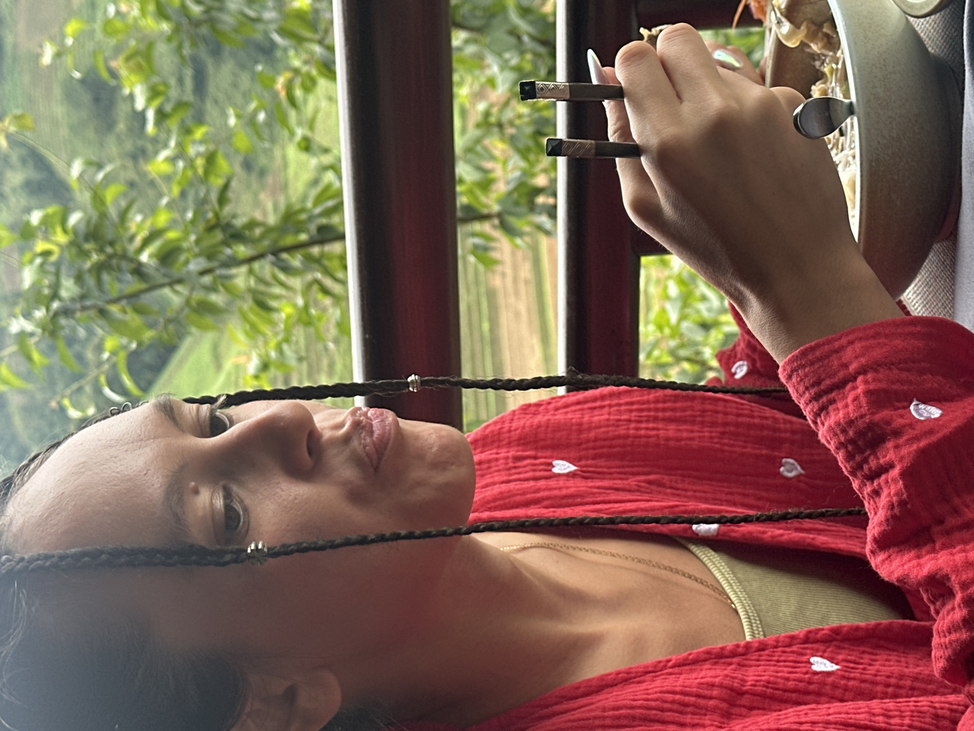 A person is seen in a close-up, three-quarter profile, sitting outdoors and eating in Púng Luông, Vietnam. The individual has dark hair styled in numerous thin braids, some adorned with small silver-colored rings or beads. Their gaze is directed slightly downwards and to the right, with lips subtly pursed as if in the act of chewing or contemplation. They are wearing a textured red garment, possibly a jacket or shirt, featuring small white embroidered hearts. A light green tank top and a thin gold chain necklace are visible underneath the red garment. The person's right hand holds a pair of dark chopsticks with patterned metallic tips, poised over a light gray or beige ceramic bowl filled with food, which appears to be a soup or noodle dish with various ingredients including what look like mushrooms or dumplings. A silver spoon is also present within the bowl. The person's fingernails are long and pointed, with an iridescent sheen. In the blurred background, lush green foliage, trees, and gently sloping hills or mountains are visible, characteristic of a natural, possibly agricultural, landscape. A dark wooden railing crosses the mid-ground, separating the dining area from the expansive view.