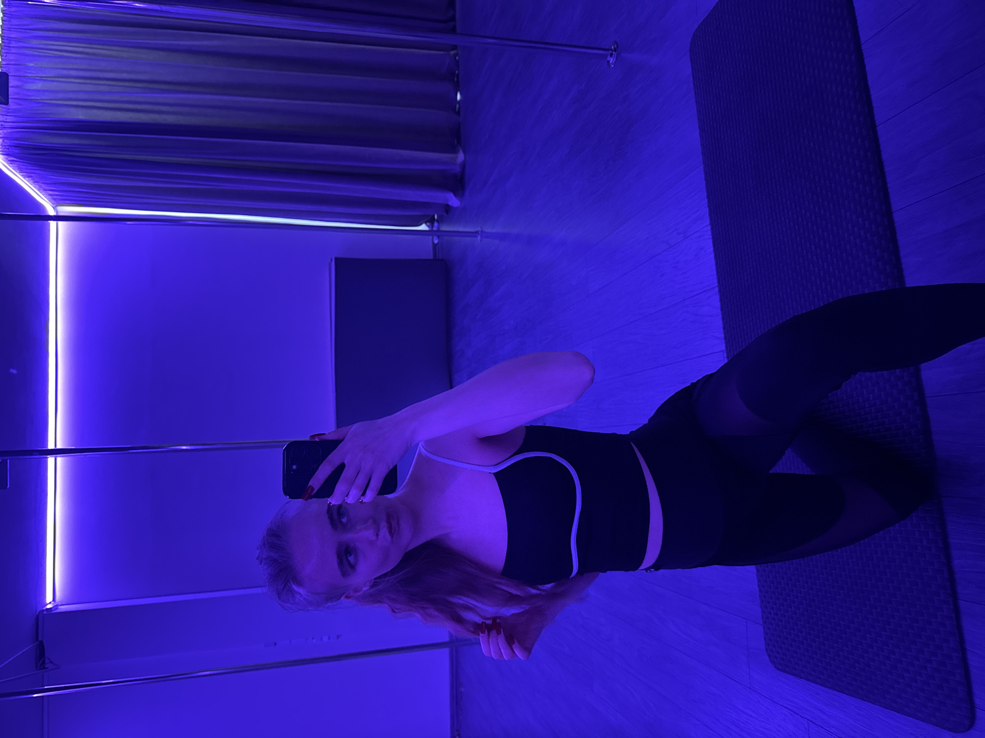 The image shows a blonde-haired woman taking a selfie in a room with blue lighting. She is wearing a black sports bra with white trim and black leggings with sheer panels. She is holding a black smartphone with manicured nails visible. The room appears to be a gym or dance studio with a pole on the left side and a grey yoga mat on the right. The floor is a light-colored wood. The presence of the yoga mat, pole and attire suggests a fitness-related activity. The city location is unknown.