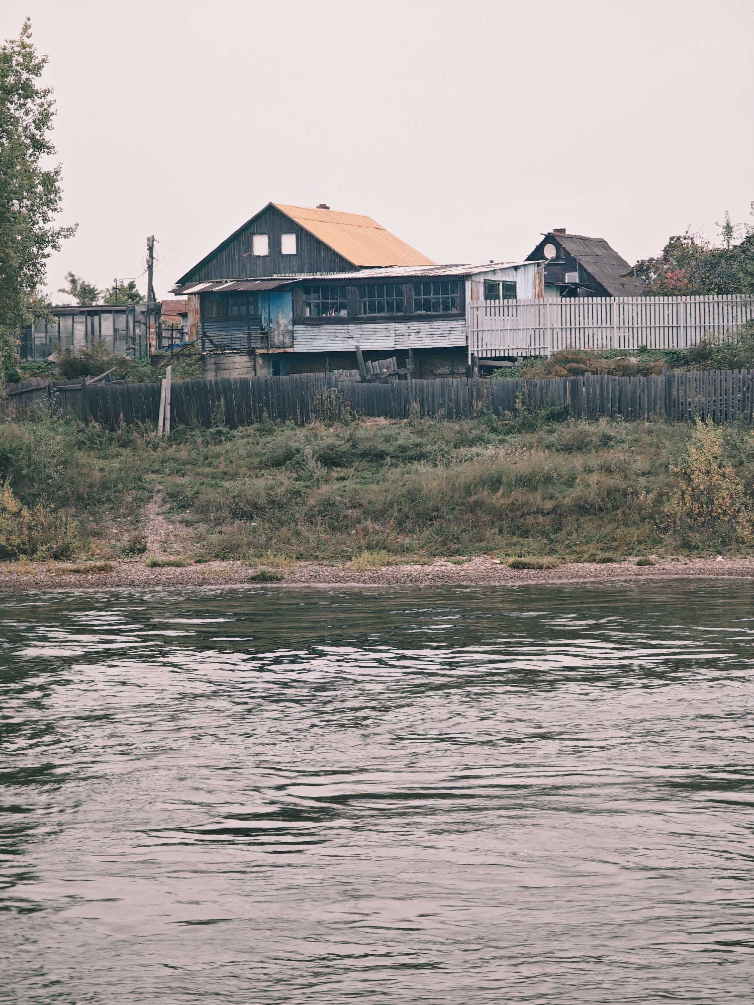 An outdoor, eye-level, wide shot depicts a cluster of rustic, weathered buildings situated along a body of water, likely a riverbank in Ermolaevo, Russia. The structures are primarily made of wood and corrugated metal, with a prominent house featuring an orange roof and a blue-painted section. A white picket fence separates the buildings from a grassy embankment covered in vegetation. The water in the foreground is calm, with visible ripples reflecting the overcast sky. The scene suggests a remote, possibly rural, setting.