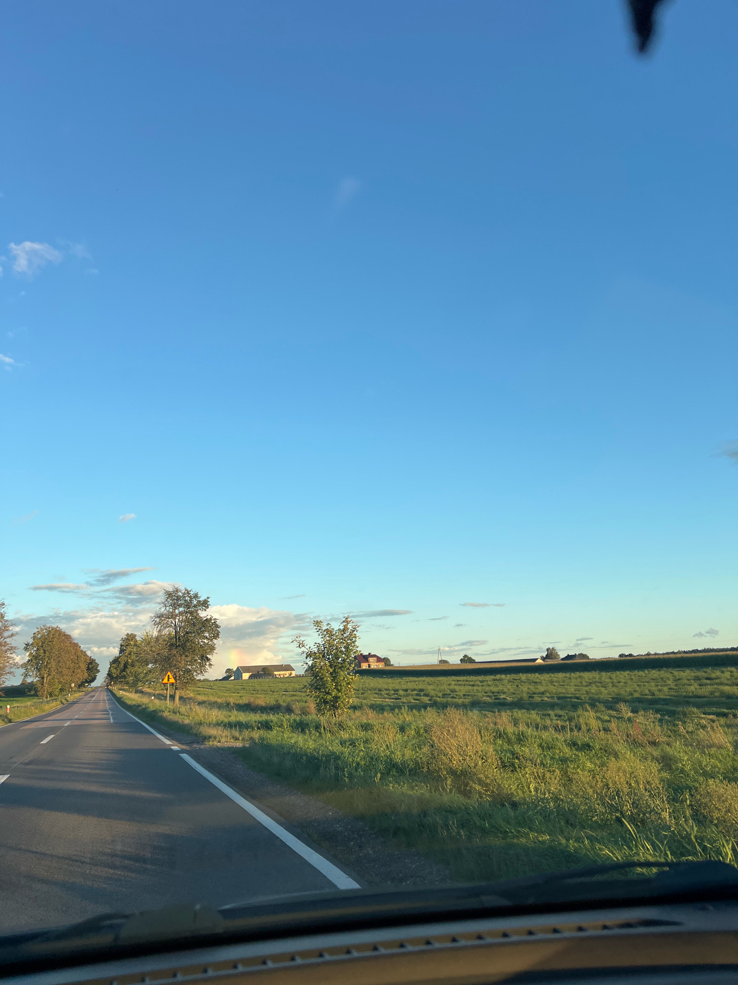 The image is a photograph taken from a vehicle's perspective, looking forward along a paved road in Kapice-Lipniki, Poland. The road, marked with white lines, curves slightly to the left and extends into the distance. On either side of the road are grassy fields, with some trees visible. In the background, there are several buildings, suggesting a rural or semi-rural setting. The sky is a clear blue with some scattered clouds, and the sun appears to be low on the horizon, casting long shadows. There is a yellow warning sign with a black symbol visible on the right side of the road. The overall impression is one of a peaceful, rural landscape under a bright sky.