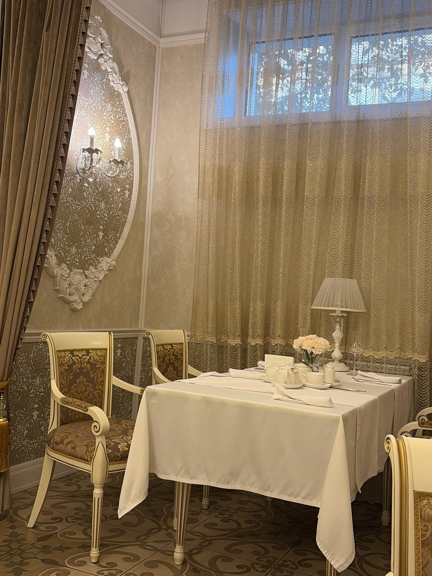 The image depicts an interior scene that appears to be a dining area in a restaurant or a formal dining room, likely located in Angarsk, Russia. The scene is set with a table covered by a white tablecloth, upon which a tea set, a vase with light pink flowers, and a table lamp are arranged. Two ornate chairs with upholstered seats and backs in a patterned fabric are positioned near the table.
The room is decorated with a damask-patterned wallpaper and features detailed white decorative molding on the wall, including a sconce with two lights. Heavy, floor-length curtains in a warm beige color are visible on the left side of the image, and sheer, gold-colored curtains with a scalloped pattern hang across a window in the background. The floor is tiled with a dark pattern. No people are present in the image. The lighting suggests an ambient interior setting, possibly during the daytime given the light coming through the window.