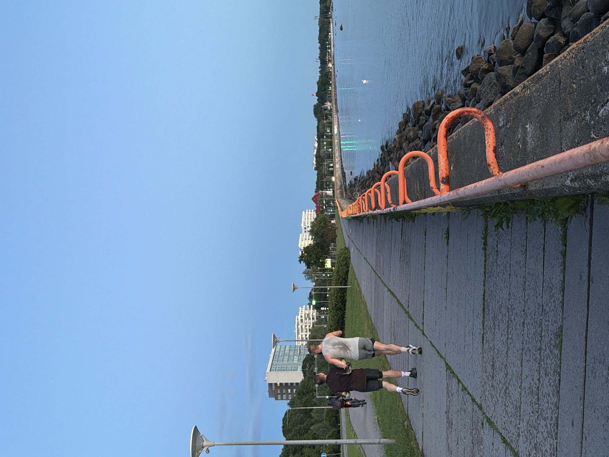 Two men are jogging on a paved pathway next to a body of water in Tallinn, Estonia. The pathway is bordered by a concrete wall with an orange railing on the right side, separating it from a rocky shoreline and the water. On the left side of the pathway, there is a grassy area with trees and streetlights. In the background, several modern buildings and some trees are visible, along with a person riding a bicycle. The sky is a clear, light blue, suggesting it is either early morning or late evening. The scene appears to be part of a waterfront promenade, likely in a developed urban area.