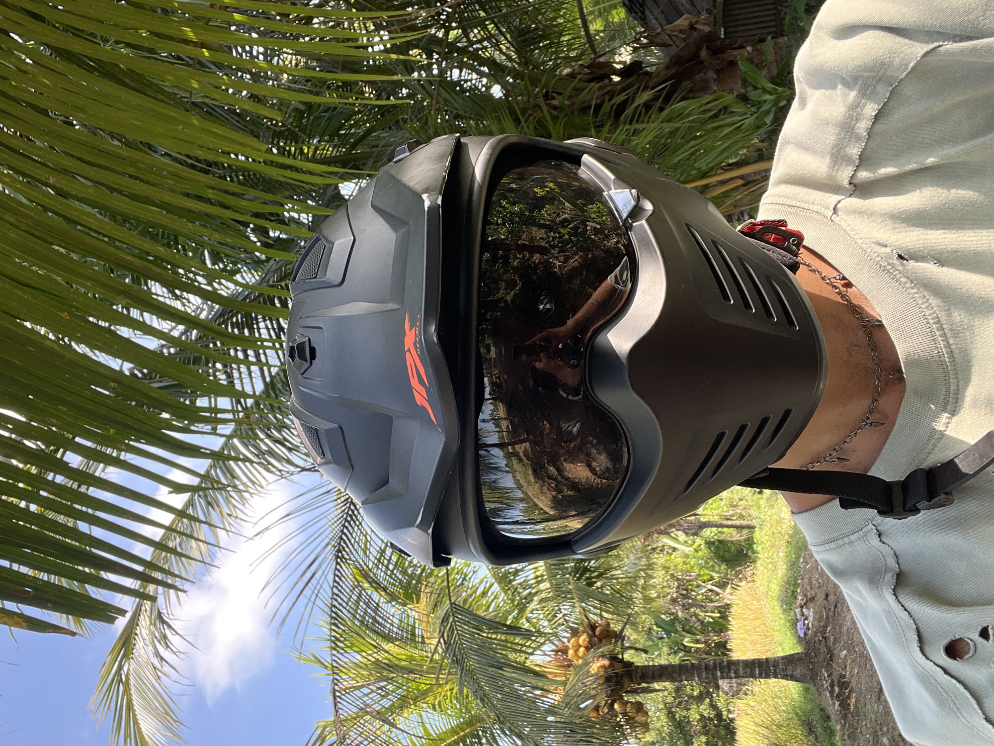 A close-up, eye-level, outdoor selfie shows a person wearing a matte black full-face motorcycle helmet with a dark tinted visor. The helmet has an orange "JX" logo on the side and ventilation grates. The visor reflects a wooded area and the sky with some clouds. The person is wearing a light olive-green t-shirt with distressed detailing around the collar and holes on the shoulder. A thin silver chain with small spike-shaped pendants is visible around their neck. The background features lush green palm fronds in the foreground, with a tropical landscape behind, including palm trees with coconuts and a rice field under a clear blue sky. This scene is located in the city of Kerambitan, Indonesia.