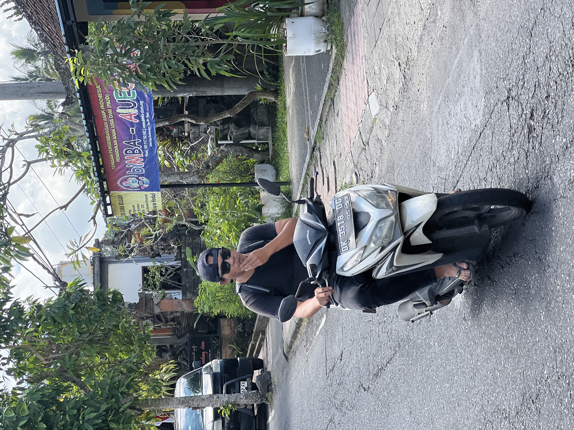 A man wearing a black t-shirt, black pants, sunglasses, and a black baseball cap is riding a white Honda scooter down a paved street in Ubud District, Indonesia. He has his right hand raised to his chin, appearing to be in thought. The scooter has a license plate that reads "DK 3518 DG." In the background, there are trees, buildings, and a banner that reads "YAYASAN PENGEMBANGAN ANAK INDONESIA PENDIDIKAN USIA DINI (PAUD) BIMBA - AIUEO." There is also a black SUV parked on the left side of the road. The day is bright and sunny.