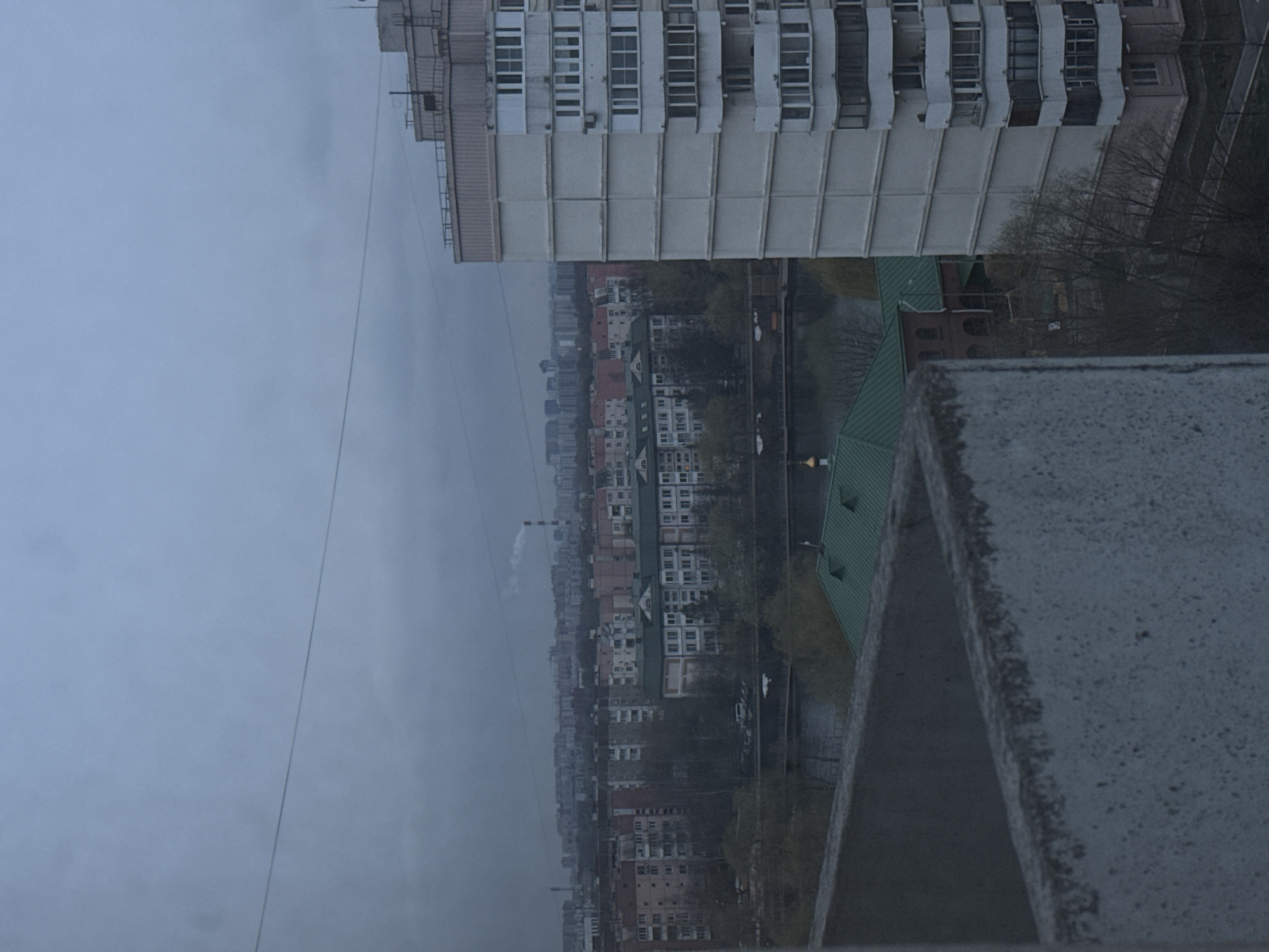The image depicts a cityscape in Moscow, Russia, under a cloudy sky. A tall, multi-story apartment building with a white facade dominates the upper portion of the view. Below, a mix of older, shorter buildings lines a road alongside a green space or park. A smokestack emits smoke into the hazy atmosphere. A concrete ledge or structure is visible in the foreground.