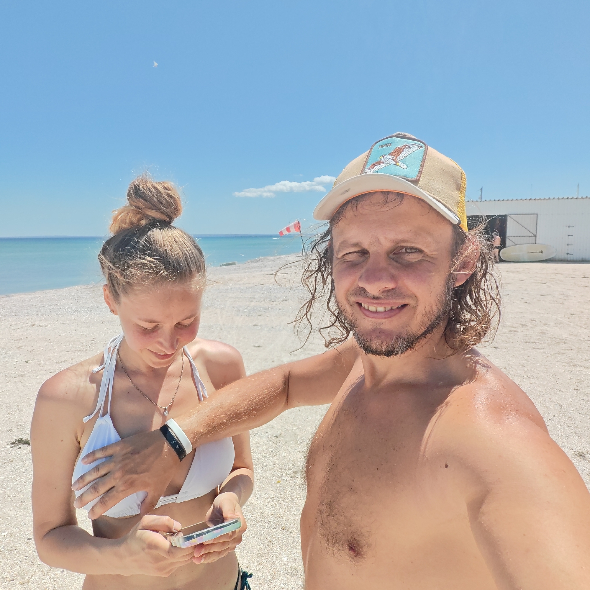 A selfie image shows two individuals, a man and a woman, on a beach. The man, who appears to be holding the camera, has long, wavy hair, a light beard, and is wearing a tan baseball cap. He is shirtless and smiling. The woman, standing to his left, is fair-skinned with her hair styled in a bun. She wears a white bikini and is looking down at a cell phone in her hand. The man's hand is on her chest.
The background consists of a sandy beach and a calm body of water, likely the ocean or a large lake, under a clear blue sky. A small white building with two paddleboards leaning against its side is visible in the distance on the right. A windsock is also visible in the middle ground.
