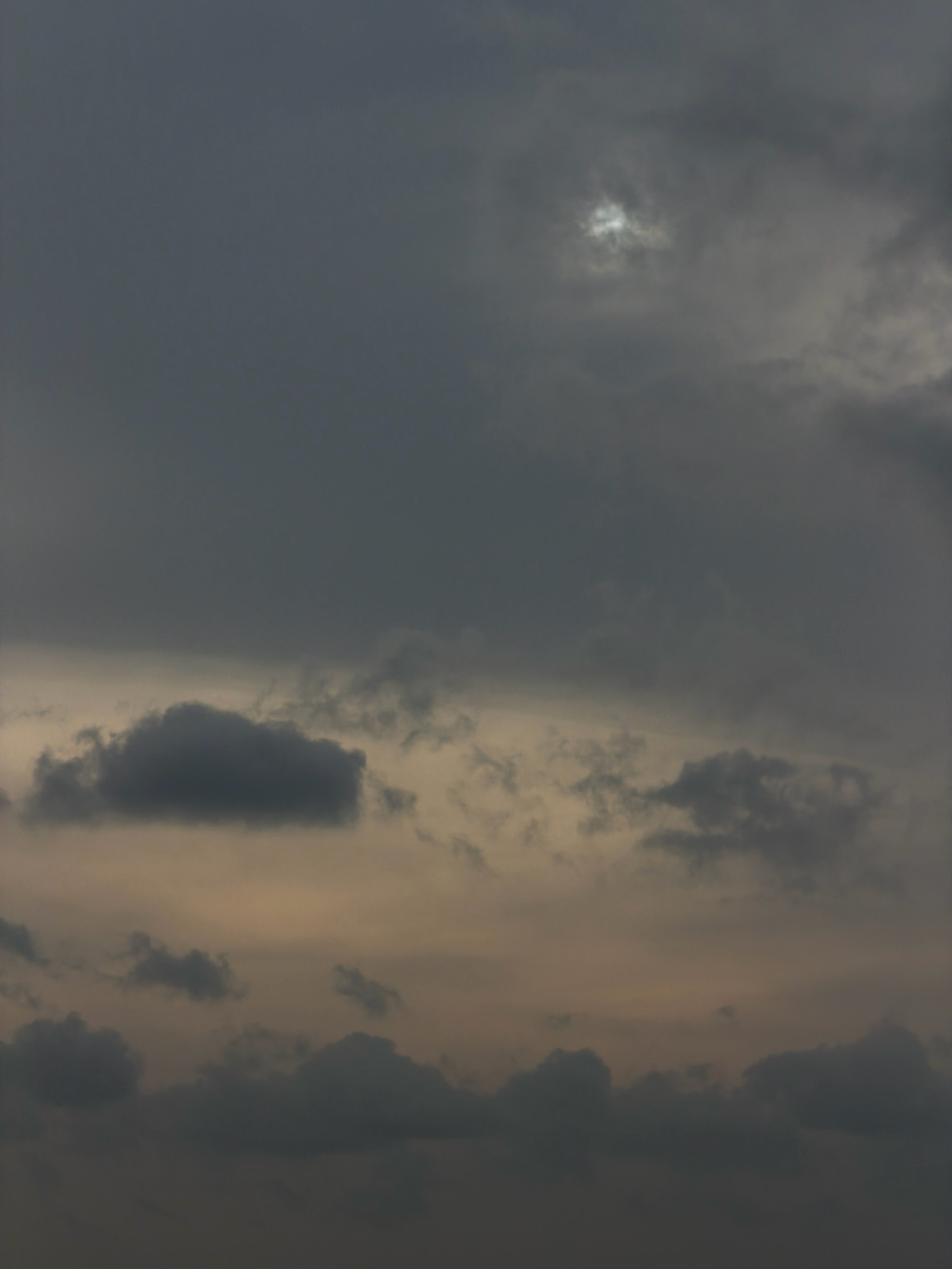 The image depicts a cloudy sky over Koorkaparamba, India. The sky is filled with dark, heavy clouds, with a small patch of brighter sky with some sunlight breaking through visible towards the top center of the image. The overall tone is muted and somber, suggesting an overcast or approaching storm. There are no people, structures, or distinct geographical features visible in this sky-only photograph. The clouds vary in density and texture, with some appearing wispy and others more solid. The lighting is diffuse due to the cloud cover.