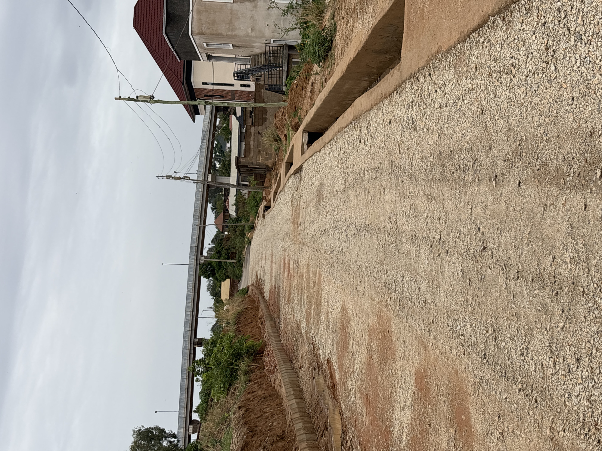 The image depicts a partially constructed road in Jos, Nigeria. The road surface is covered in loose gravel and reddish-brown soil. A concrete drainage channel runs along the upper right side of the road. A pedestrian bridge crosses above the road in the distance. In the background, houses and utility poles are visible, with an overcast sky overhead. The foreground shows newly exposed earth along the sides of the road, indicating recent construction activity. No people are visible in the frame.
