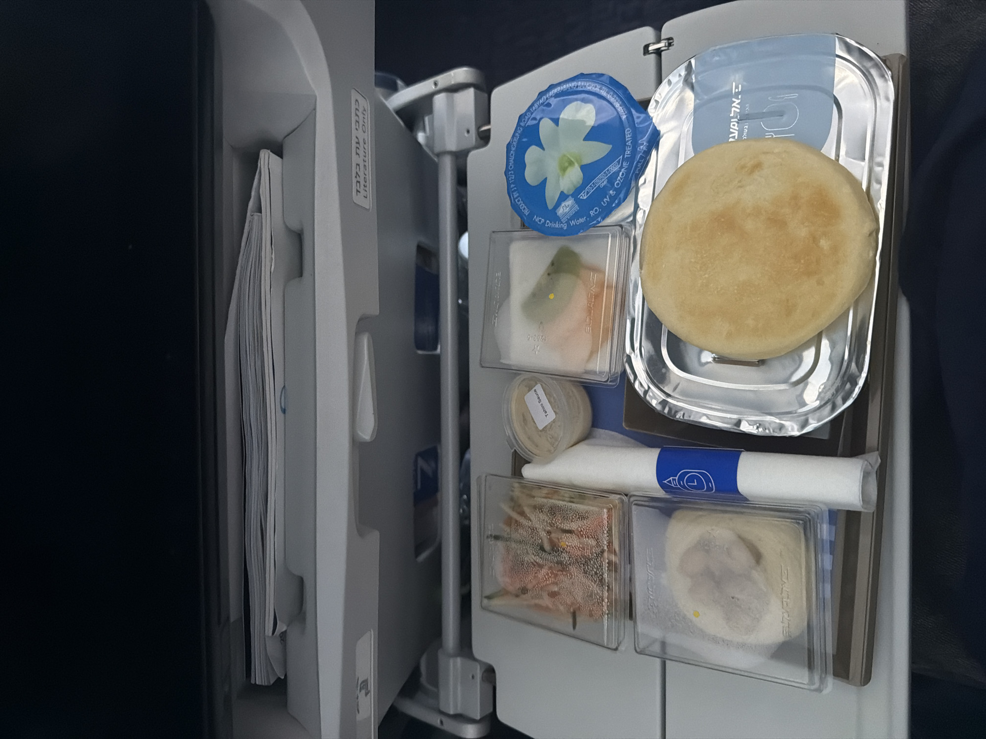 The image shows a tray of food on an airplane. The tray includes a round flatbread, a container of water, a container of tahini sauce, and several other small containers with food items in them. There is also a rolled napkin. The tray is attached to the back of an airplane seat. A label on the seat is written in Hebrew and English.