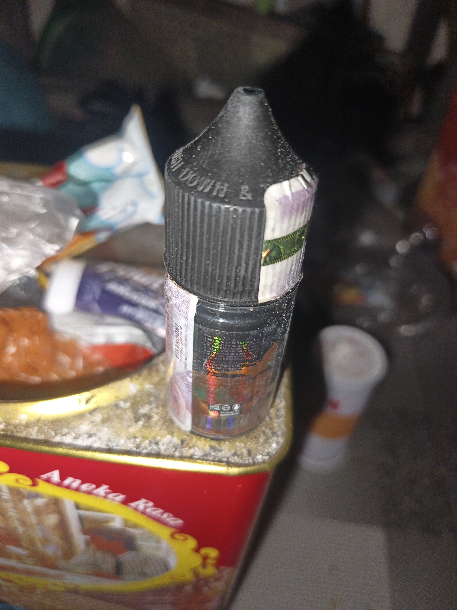 In a cluttered, indoor environment, possibly a workshop or storage area, a clear plastic bottle containing e-liquid is positioned on a textured surface. The bottle has a black cap with ribbed detailing and a warning label. Behind the bottle, several out-of-focus items are visible, including what appears to be food packaging and other containers. A red and yellow tin with the inscription "Aneka Rasa" is prominent in the foreground. The overall lighting is dim, with some areas brightly illuminated, suggesting the use of artificial light. The scene is located in Temuwulan, Indonesia, implied by the "Aneka Rasa" branding, which is a well-known Indonesian food brand, and likely indicates the origin of the tin.