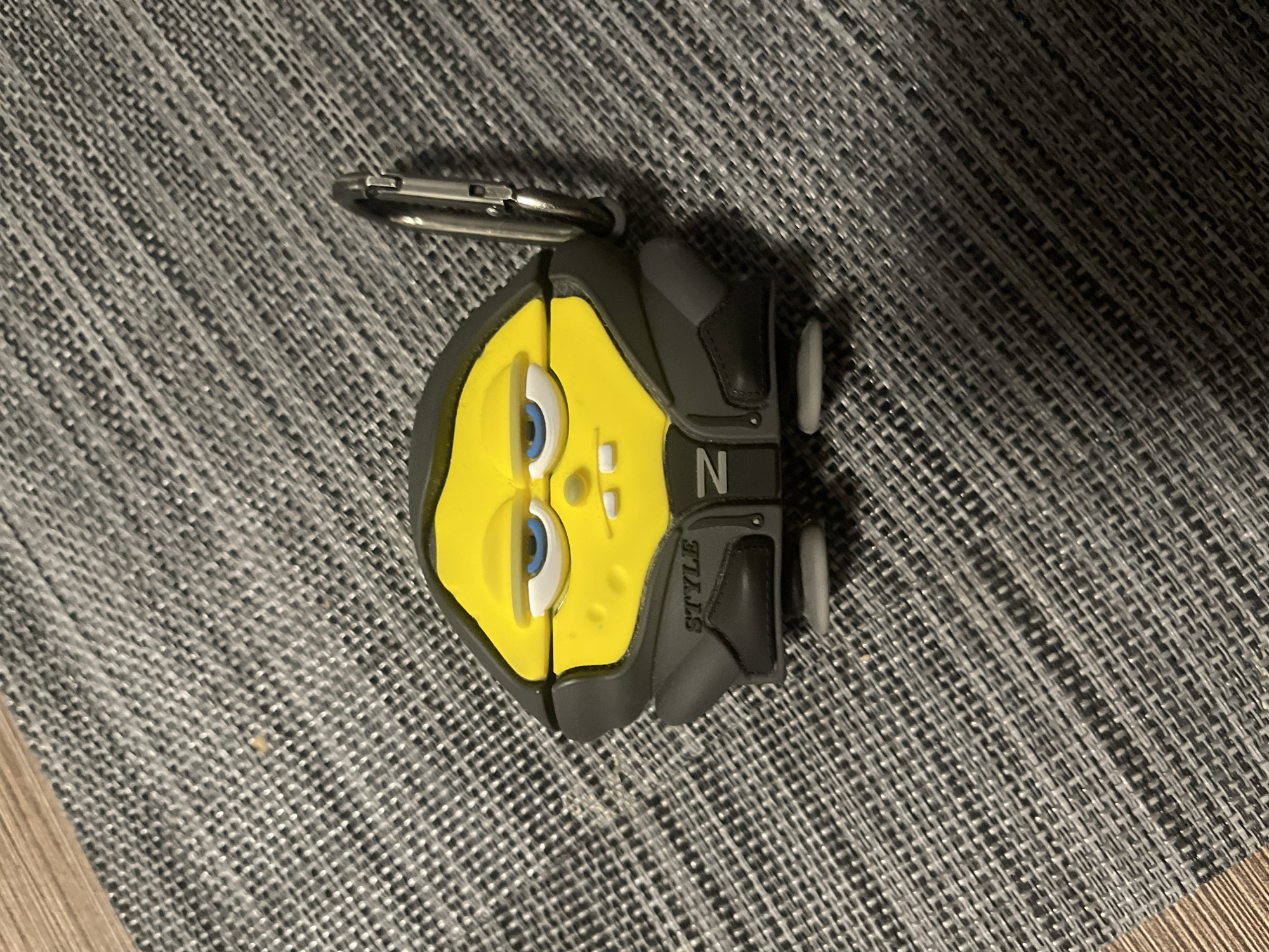 A yellow, Spongebob-shaped keychain holder is positioned on a grey, textured surface. The holder has a black jacket design with the word "STYLE" and the letter "N" visible. A silver carabiner is attached to the top left of the holder. The background is a close-up of a woven mat with wooden flooring visible in the top left corner. The image was taken in Ząbki, Poland.