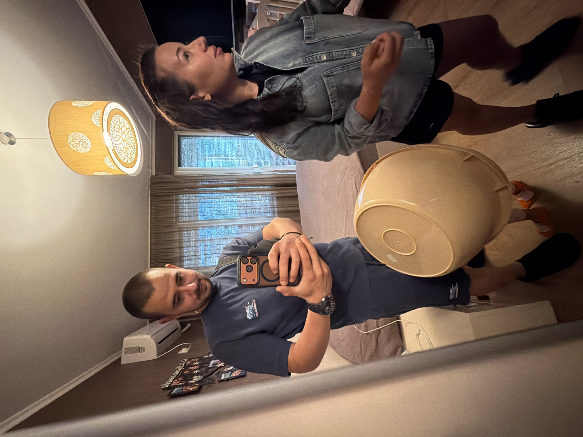 The image depicts an indoor scene, likely a bedroom, with a man and a woman. The man is in the foreground, taking a selfie in a mirror. He is wearing a navy blue t-shirt and shorts, with a watch on his left wrist. He is holding a smartphone in his right hand, and a beige bucket is partially visible in front of him. The woman is standing to his right, looking away from the camera. She is wearing a denim jacket over a black t-shirt and dark shorts.
The room appears to be furnished with a bed, air conditioning unit, and decorative items on the wall. A patterned lampshade hangs from the ceiling. The overall impression is a casual, domestic setting. The provided information about Burgas, Bulgaria, does not appear to be directly inferable from the visual elements of the image.