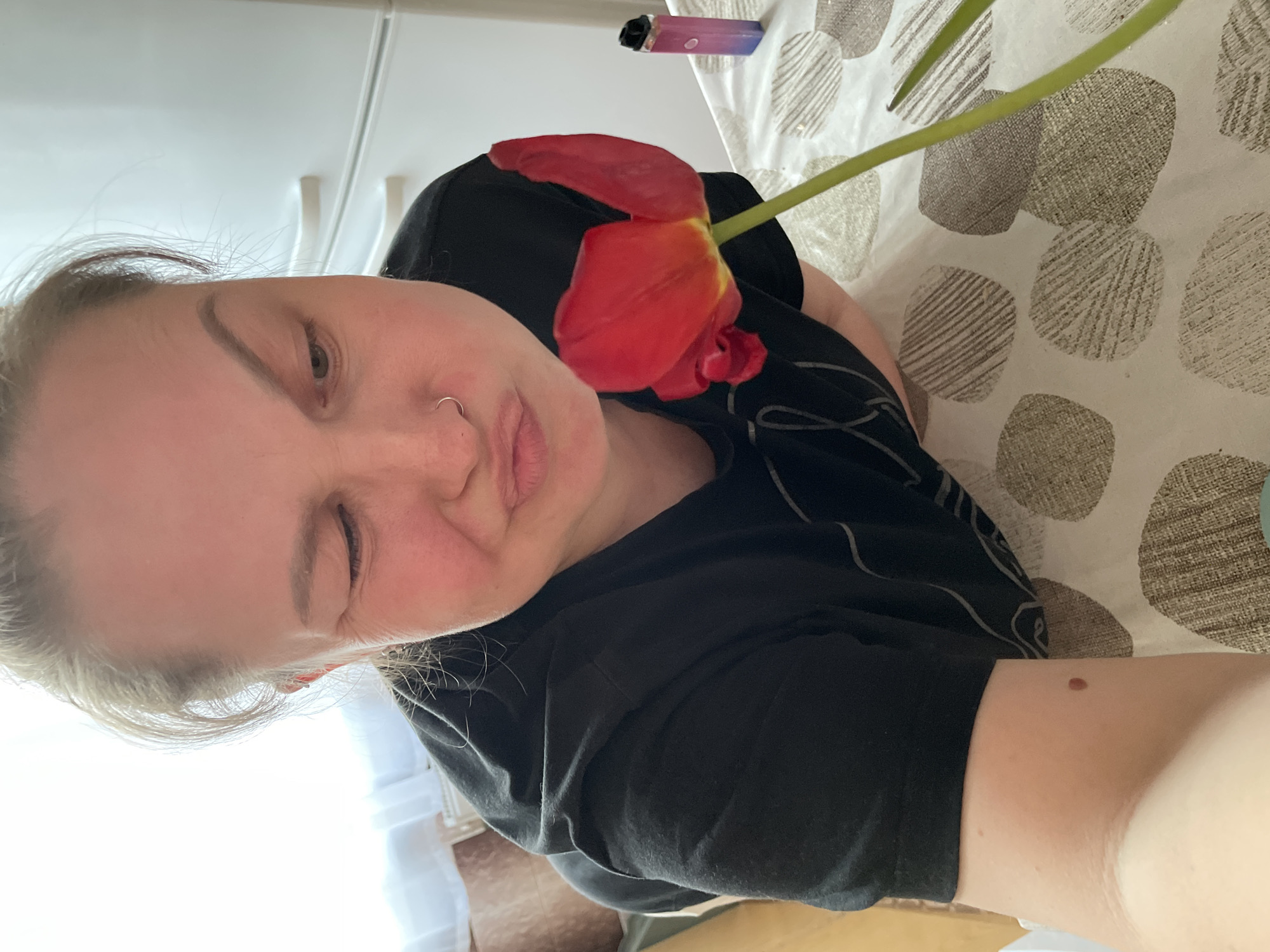 A person is pictured in a selfie-style image, with a red tulip placed near their face. The individual has fair skin and is wearing a black shirt. A nose ring is visible. The background includes a table with a patterned tablecloth and a partially visible vape pen. There's no indication that the image was taken in Izhevsk, Russia.
