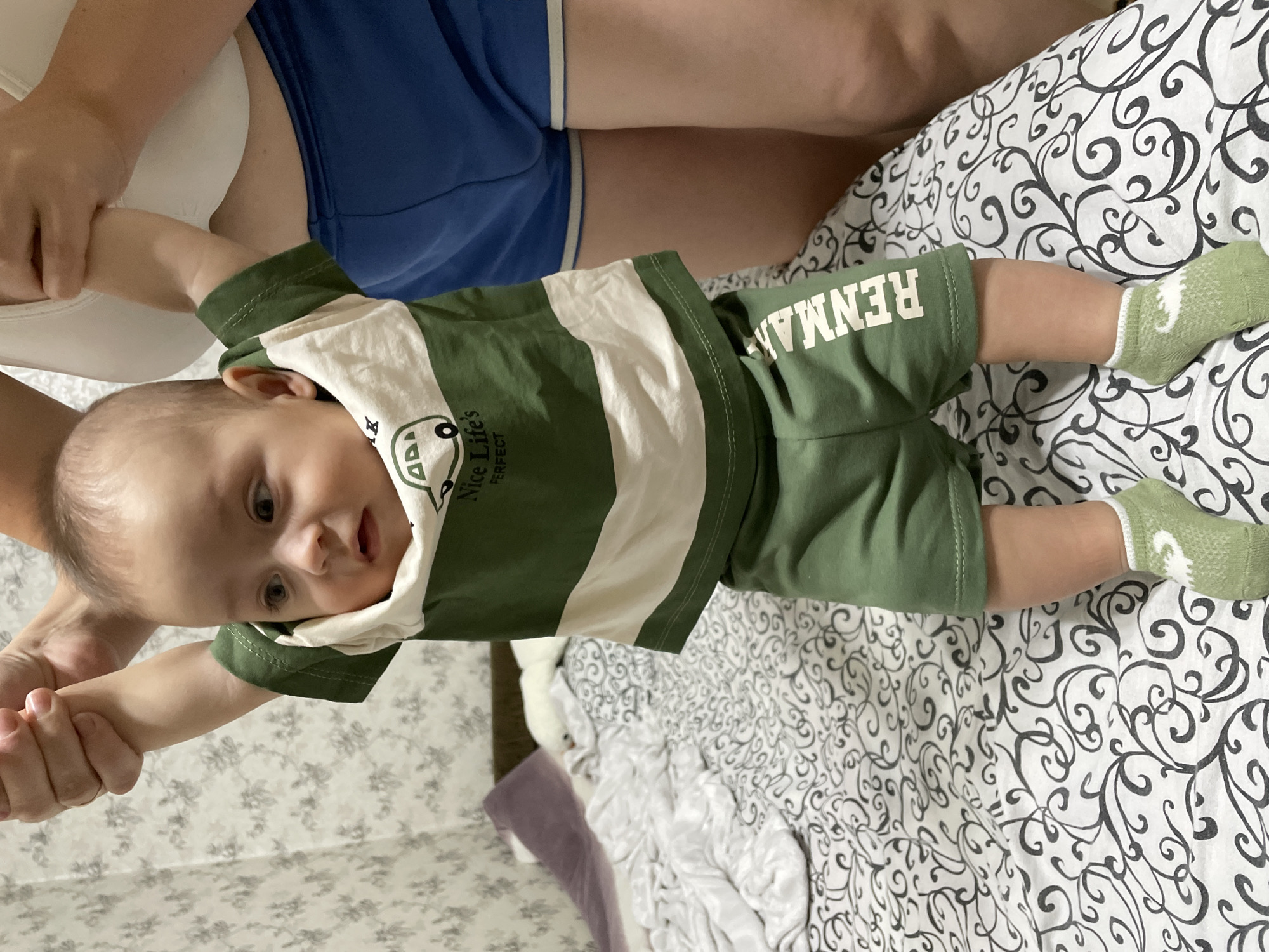 The image shows a baby standing on a bed, being held by unseen adults. The baby is wearing a green and white striped t-shirt with a picture of a bus and the text "Nice Life's Perfect", green shorts with white text that reads "RENMAN", and light green socks with a white flamingo pattern. The bed has a white and black patterned comforter. The background shows a wall with floral wallpaper and a portion of a person wearing blue shorts and a white top. The scene appears to be indoors, likely a bedroom. The location in Fălești, Moldova is not discernible from the image itself.