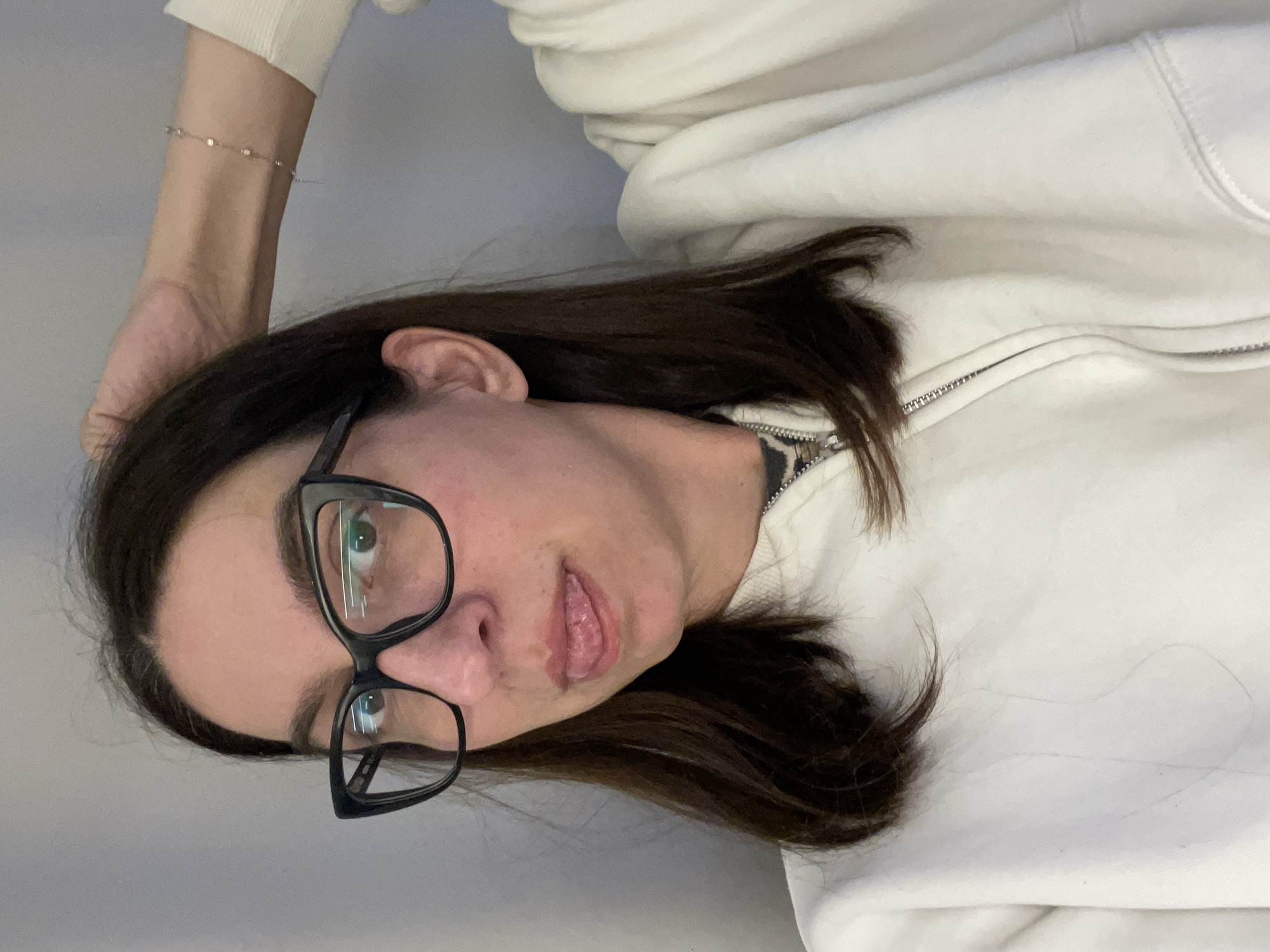 A woman with dark hair and glasses is wearing a white hoodie. She has a small bracelet on her left wrist. The background is a light grey wall. There is no information to determine the location as Sofia, Bulgaria, from the image.