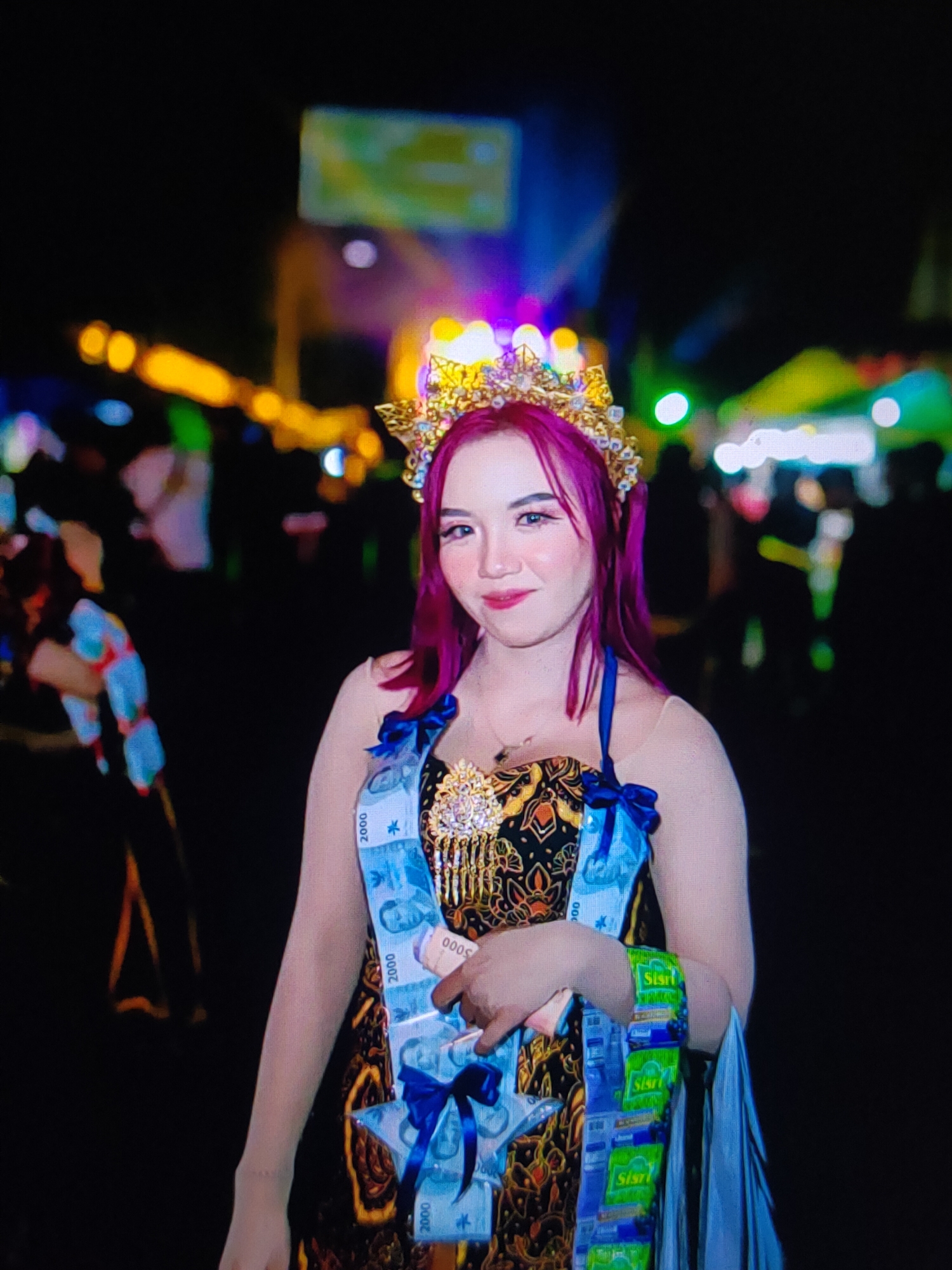 A young woman with vibrant purple hair and a golden crown poses for a photograph at night. She is adorned with a sash made of Indonesian Rupiah banknotes, and a decorative brooch on her chest. She wears a traditional patterned dress and blue ribbons. In her right hand, she holds a small stack of pink paper. The background is blurred and illuminated by colorful lights, suggesting an outdoor event or festival. Several people are visible in the background, also blurred by the low light conditions. The scene is captured at an outdoor event in an unspecified urban location at night.