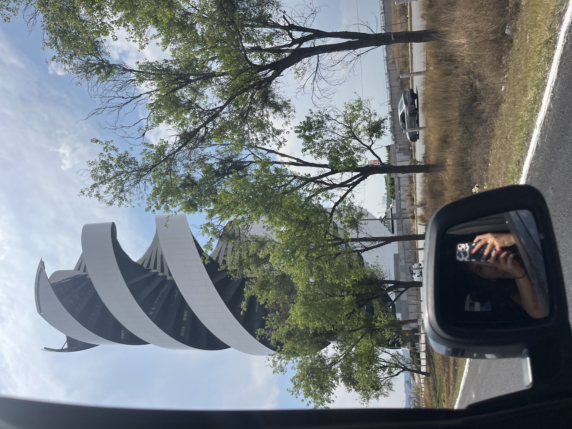 A passenger in a vehicle traveling on a road captures an image featuring the side view mirror reflecting the photographer taking the picture, along with the surroundings. The exterior shows a modern architectural structure with spiraling white elements, trees, a fence, and sparse vegetation next to the road. A parked car is visible further away. The sky is partly cloudy. The location is San Bernardino Tlaxcalancingo, Mexico.